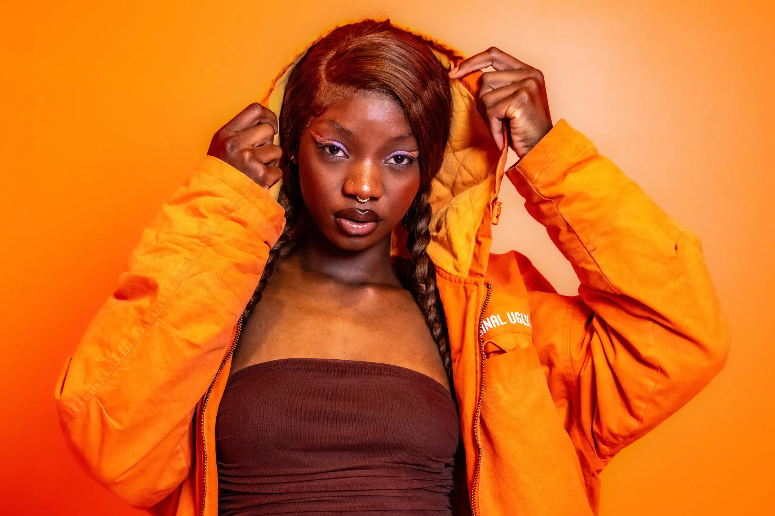 A woman with dark skin and braided hair, wearing an orange jacket and a dark top, poses against an orange background.