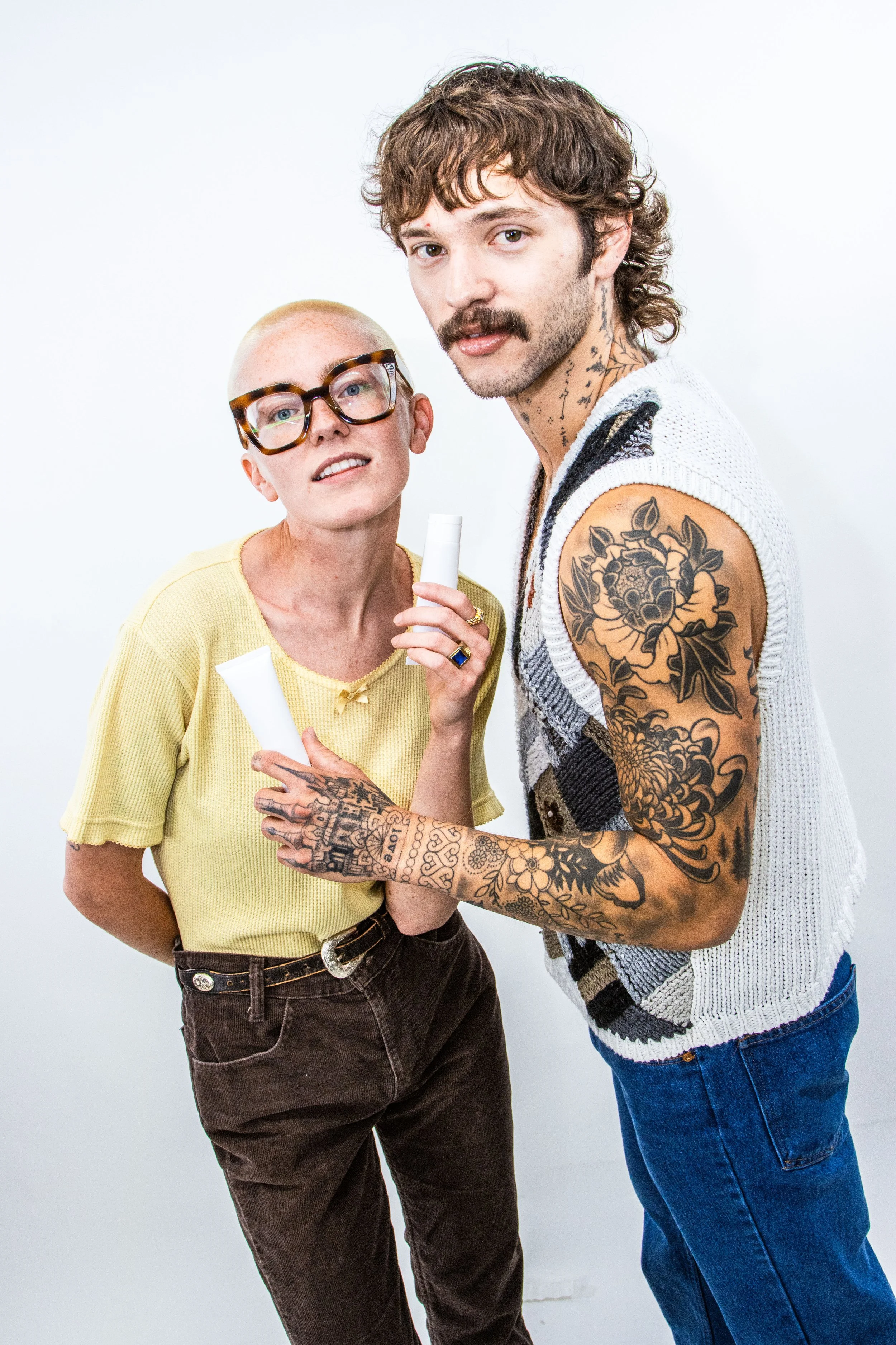 A man with brown, curly hair and tattoos on his arms and neck stands next to a woman with short, blonde hair, glasses, and a yellow shirt. They are holding tubes with a white background.