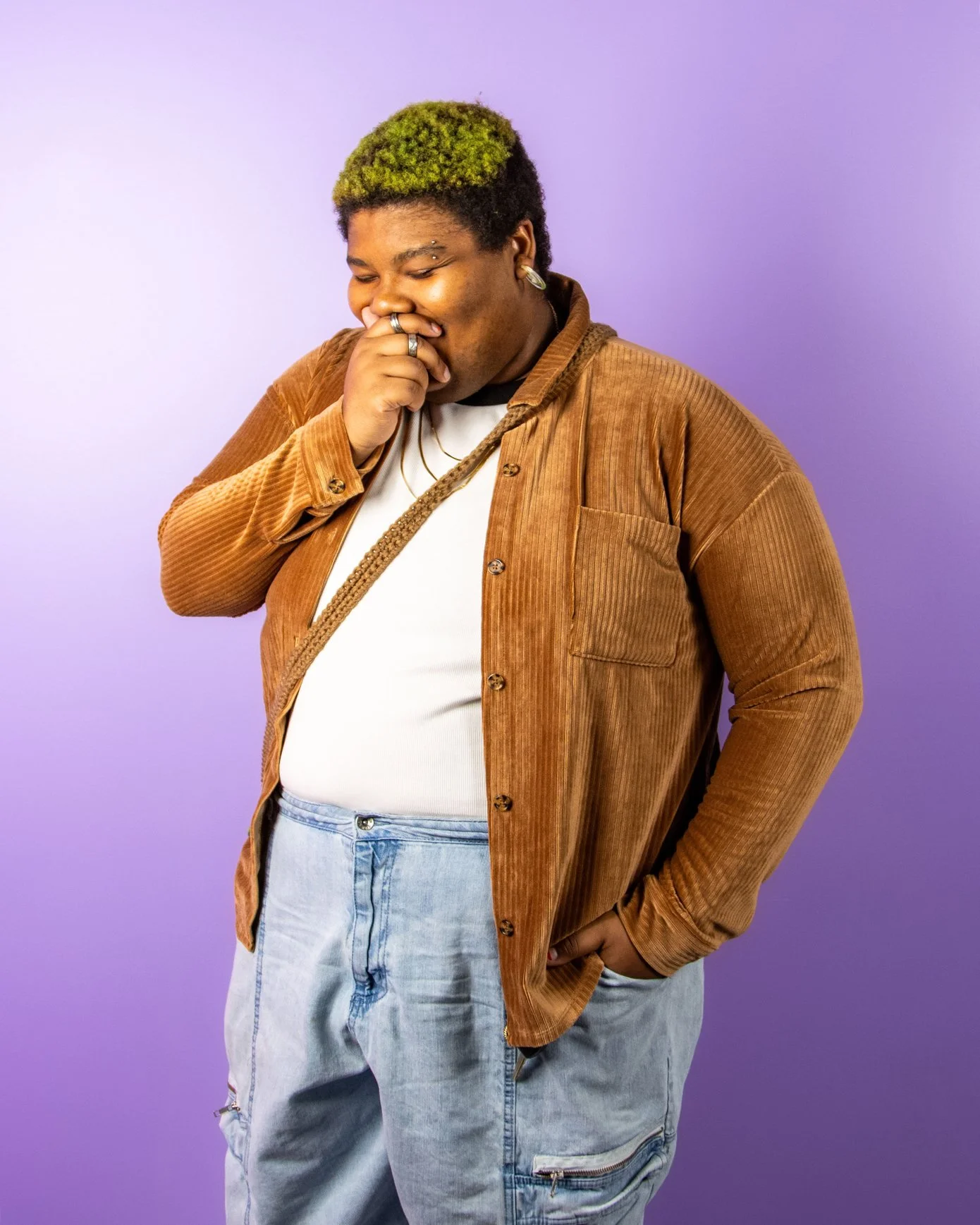 A person with green-dyed hair, wearing a brown corduroy jacket, white shirt, and light blue jeans, standing against a purple background, smiling while covering their mouth with one hand.