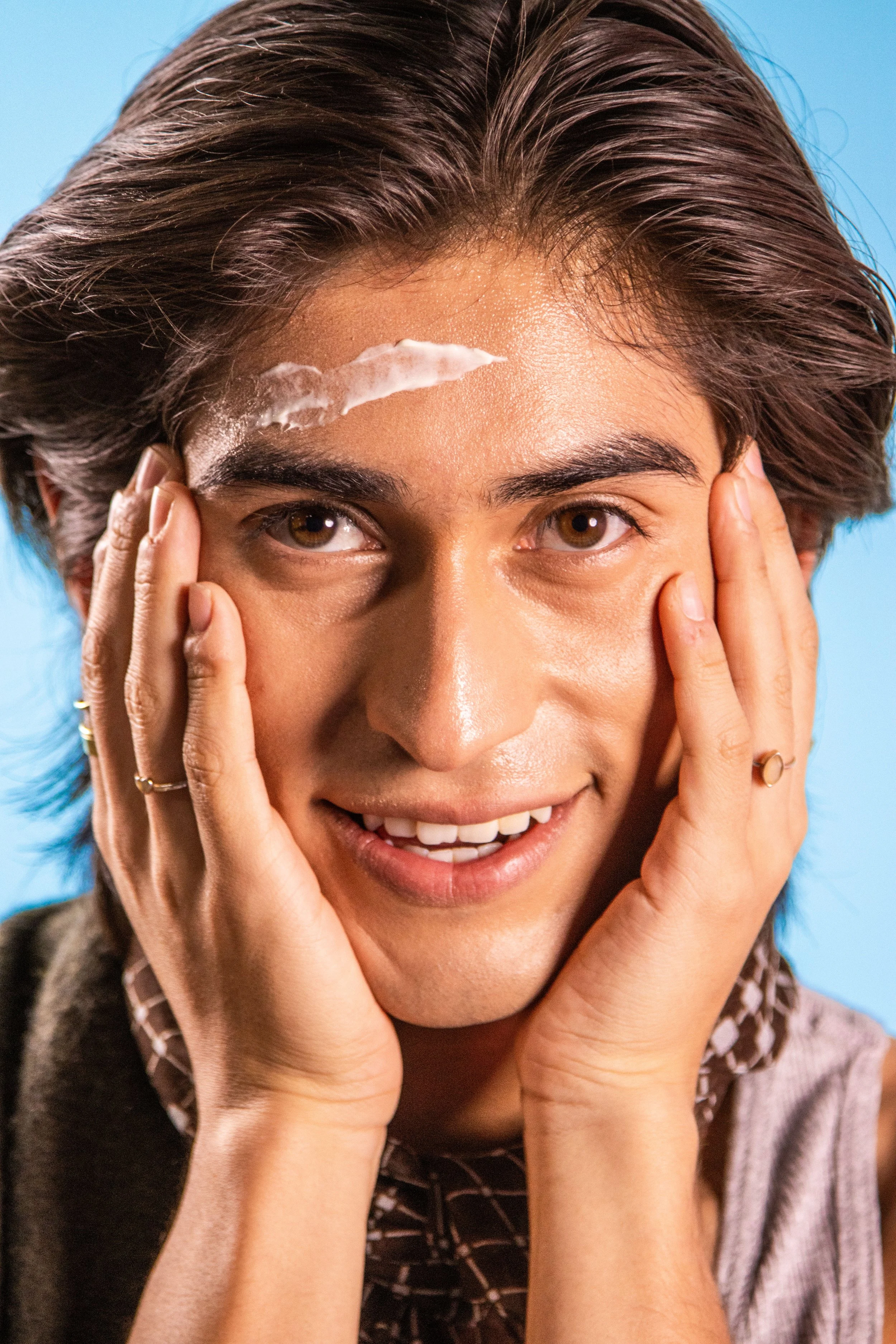 A person smiling, touching their face with both hands, with a smear of cream or lotion on their forehead. They have short, brown hair and are wearing rings.