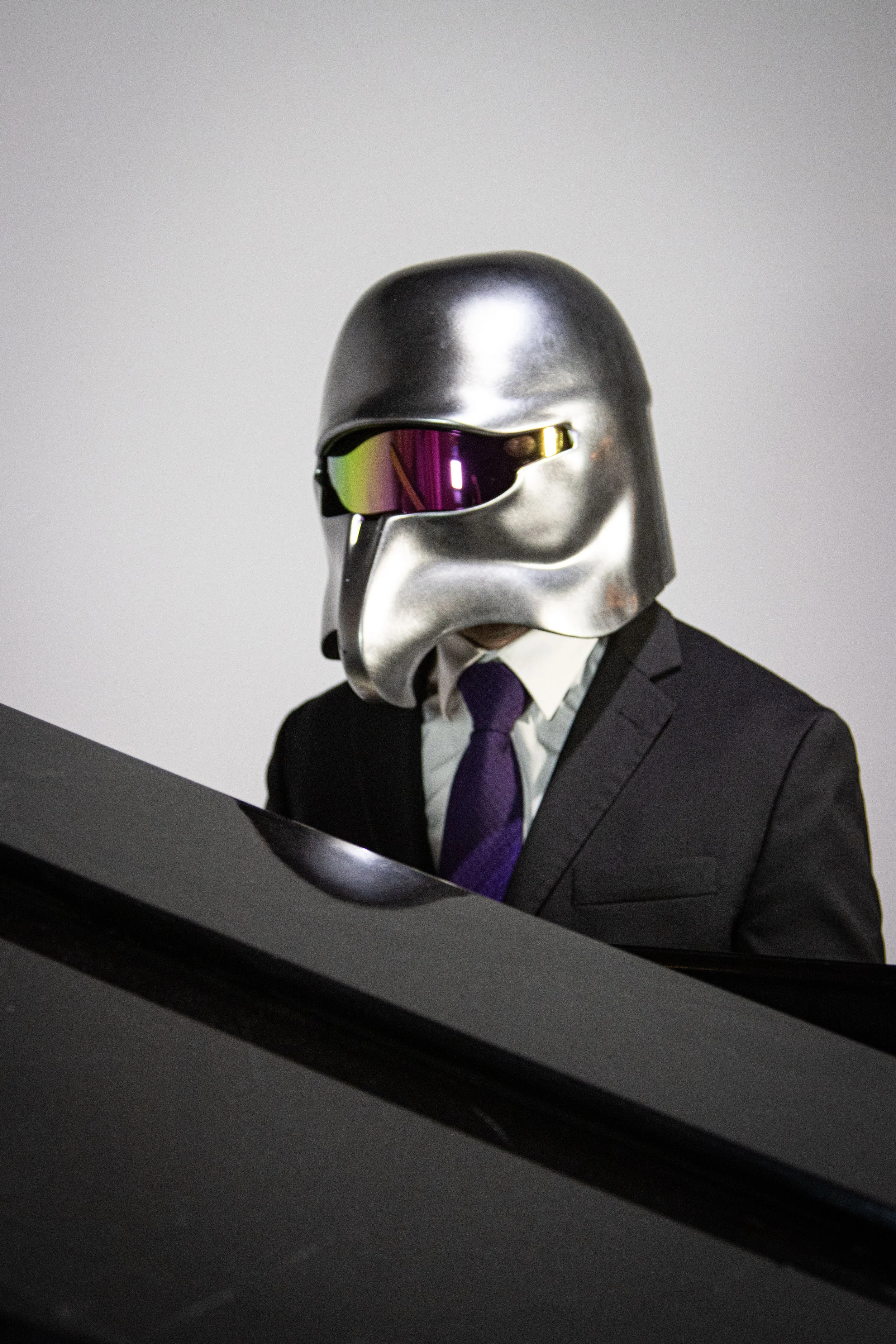 A person dressed in a business suit with a tie, wearing a metallic helmet with a futuristic visor, sitting at a desk with a computer.