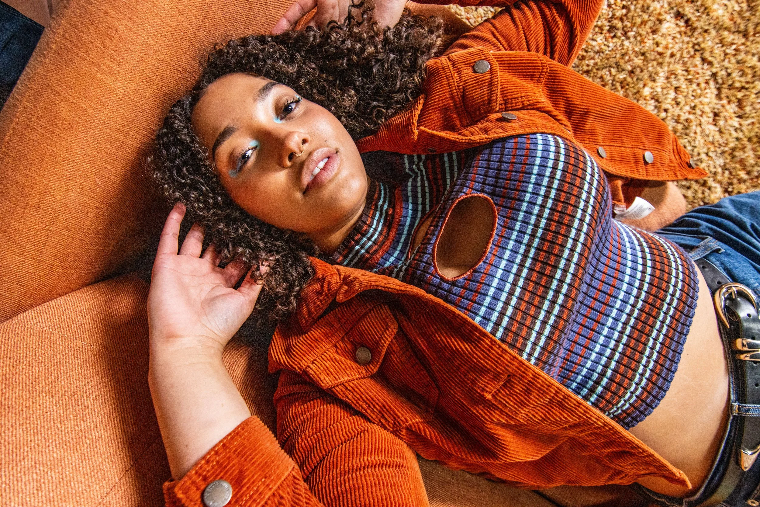 A woman with curly hair lying on an orange couch, wearing a plaid shirt with a cutout and orange jacket, looking at the camera.