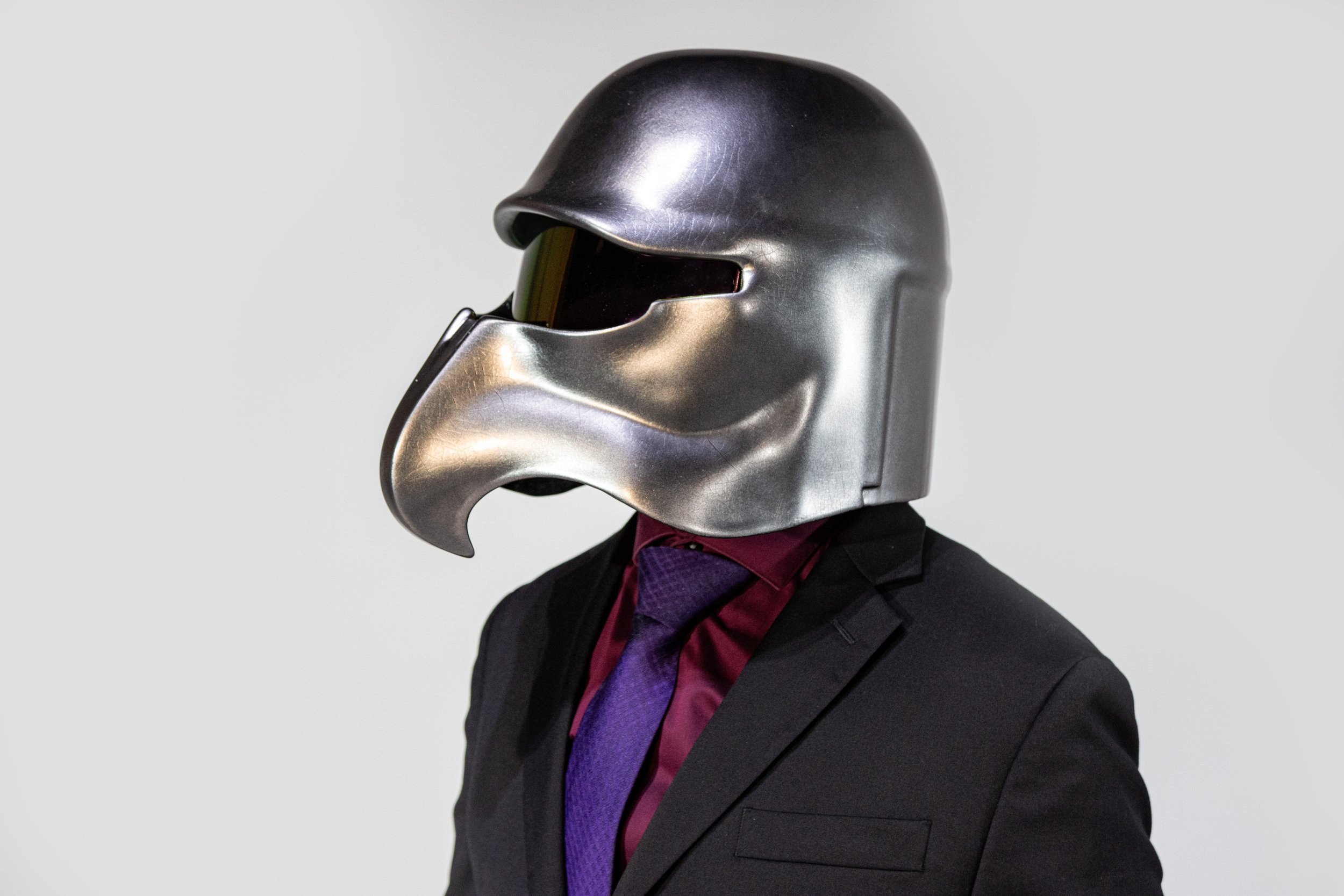 A person wearing a metallic helmet that resembles a robotic or futuristic design, dressed in a black suit with a dark purple shirt and tie, against a plain gray background.