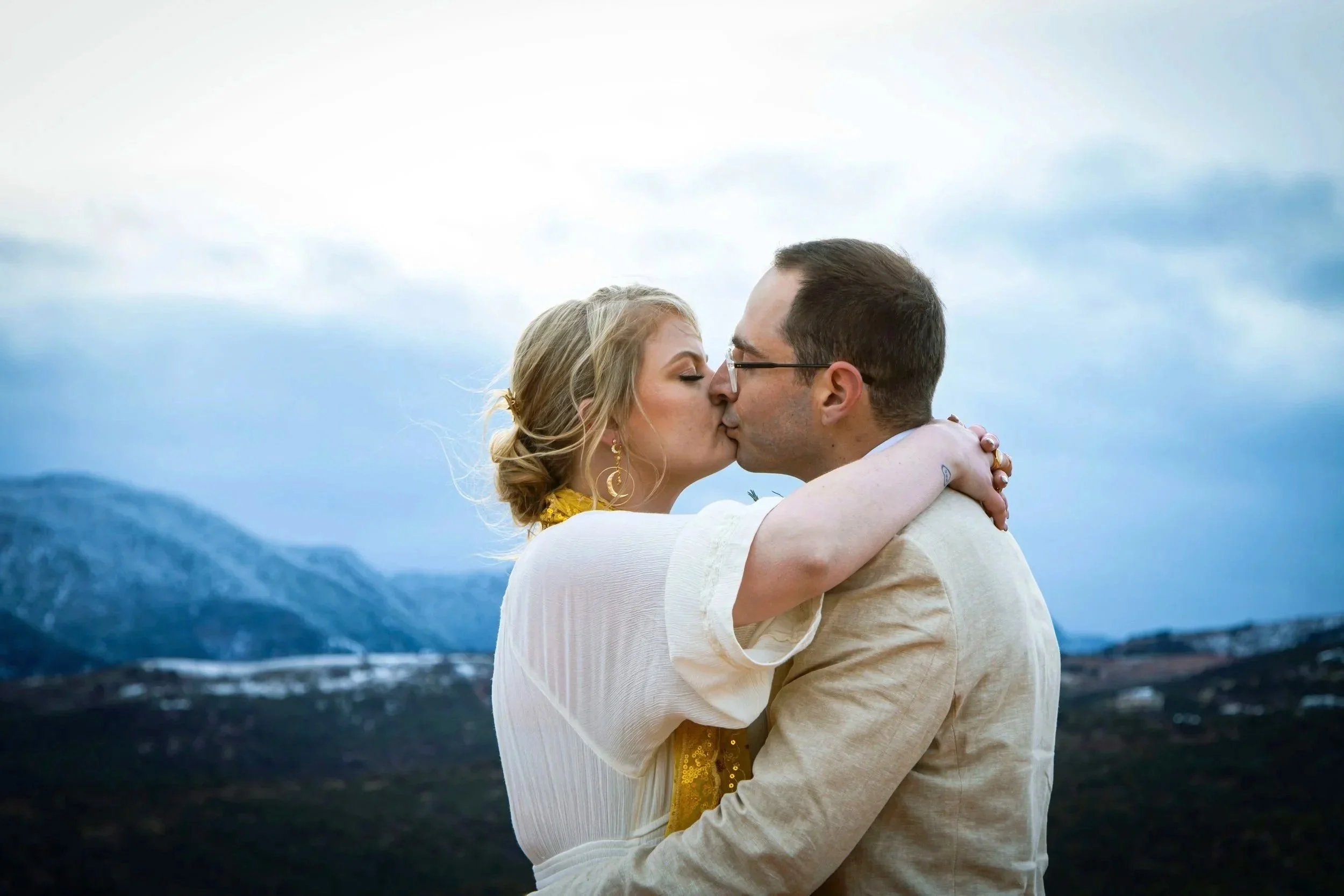 A couple sharing a kiss outdoors with mountains and cloudy sky in the background.