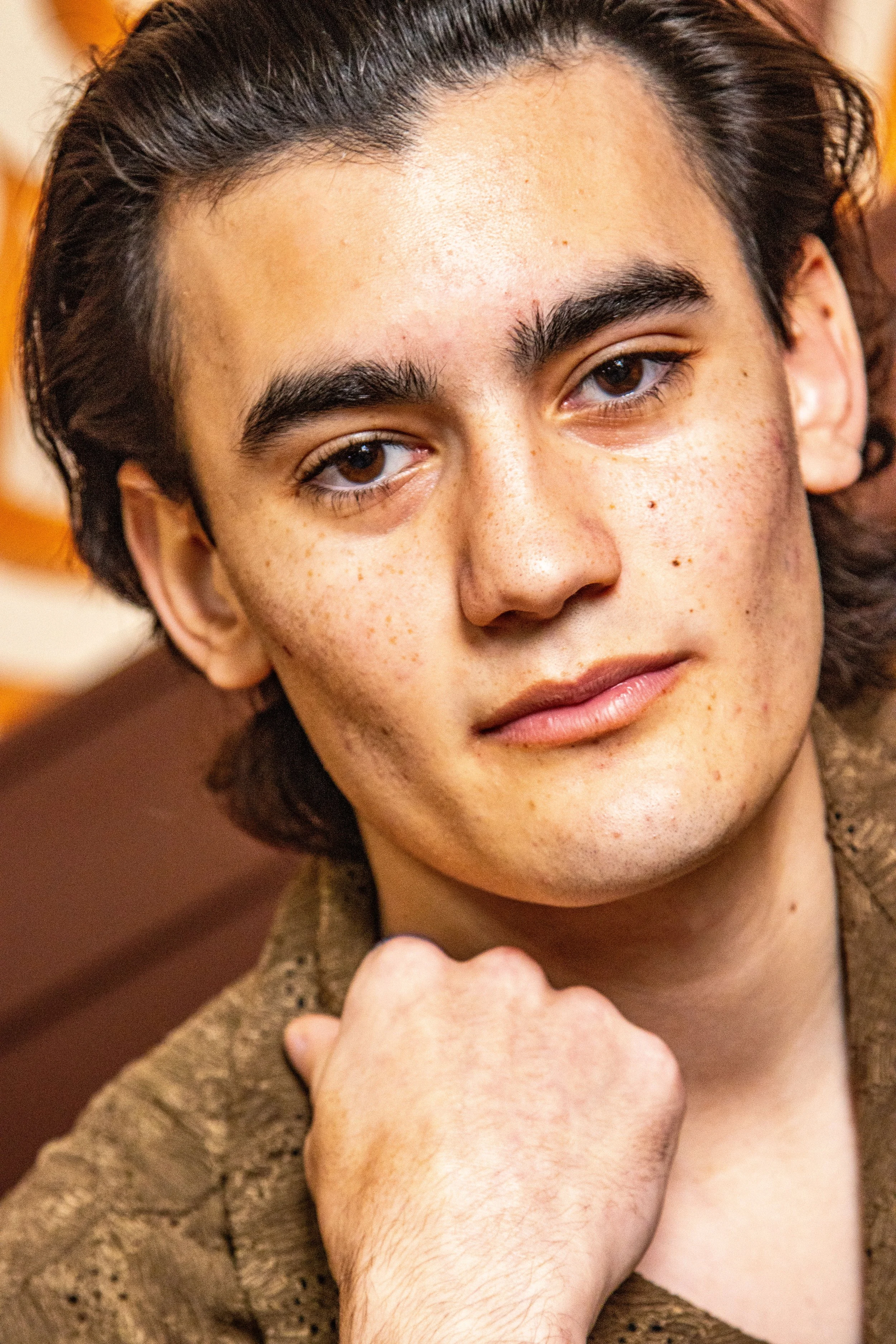 Close-up of a young man with brown eyes, dark hair, and freckles, looking at the camera with a serious expression, resting his chin on his hand.