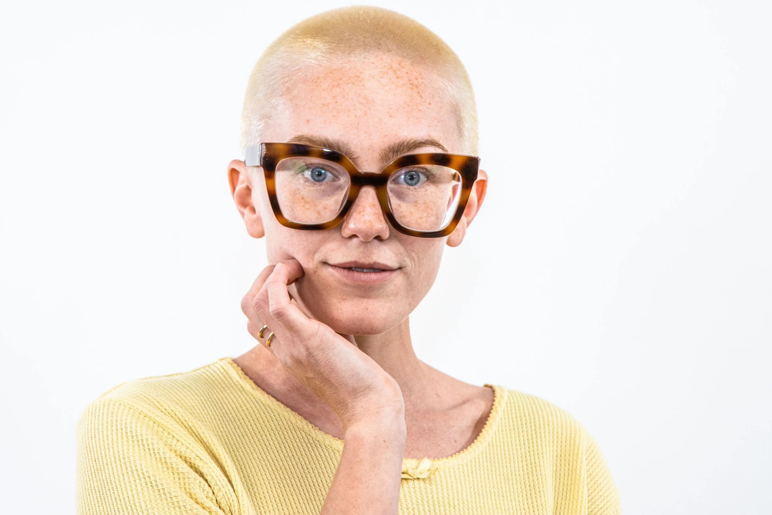 A woman with short blond hair, wearing big tortoise shell glasses and a yellow top, touches her chin with her finger, standing against a white background.