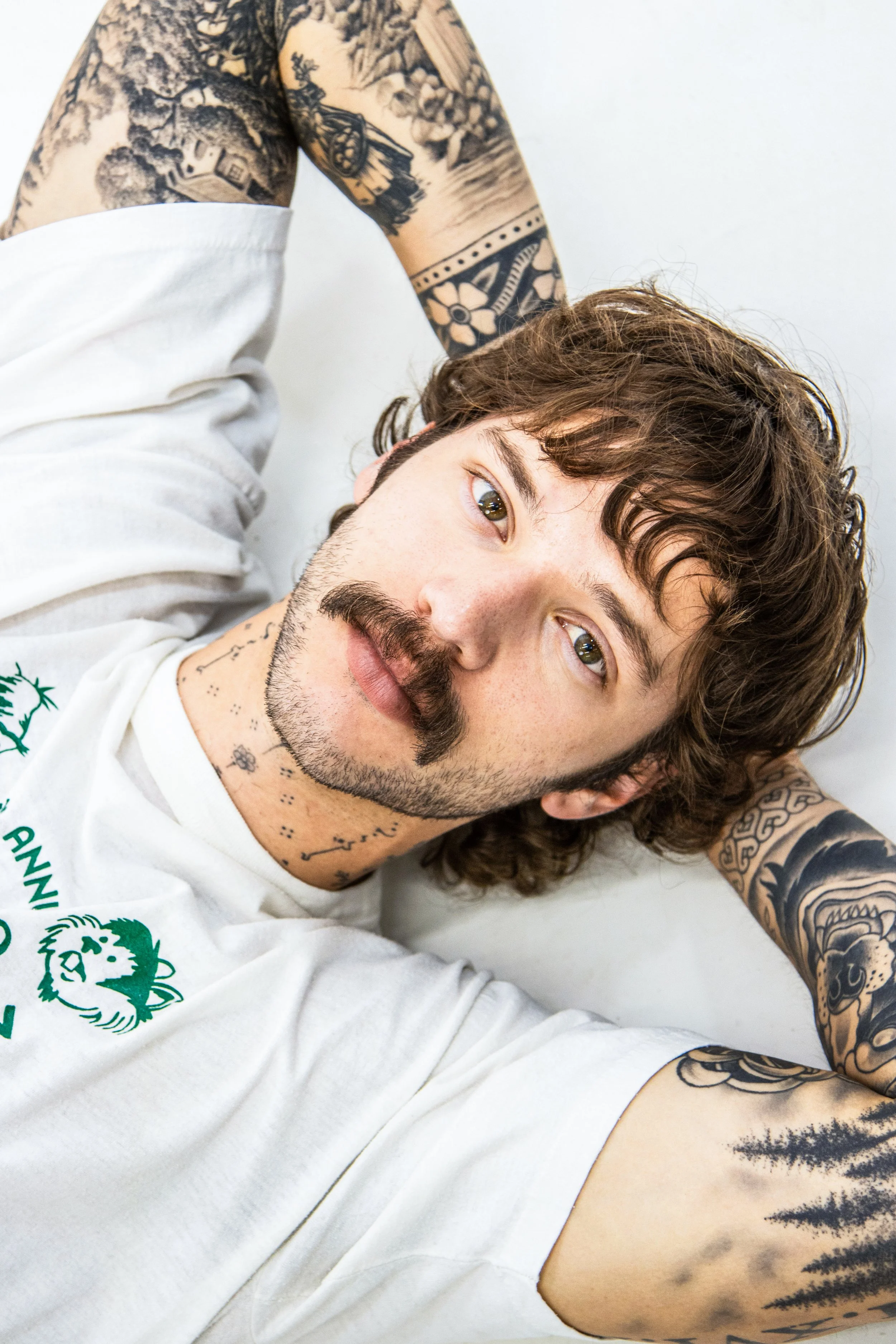 A man lying down, smiling, with brown curly hair and tattoos on his arms and neck, wearing a white shirt with green text and a lion logo.
