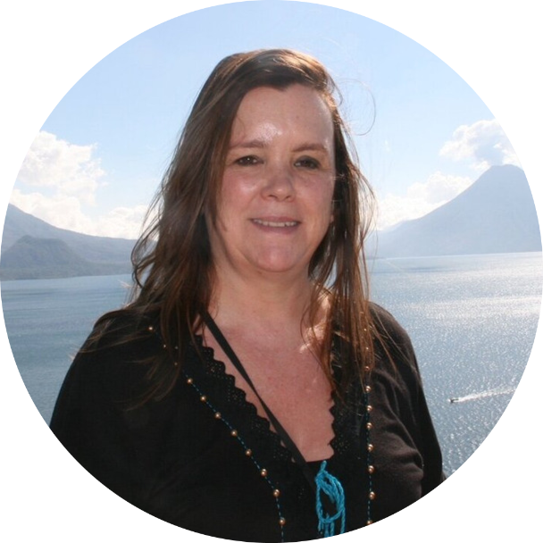 A woman smiling outdoors with a body of water and mountains in the background.