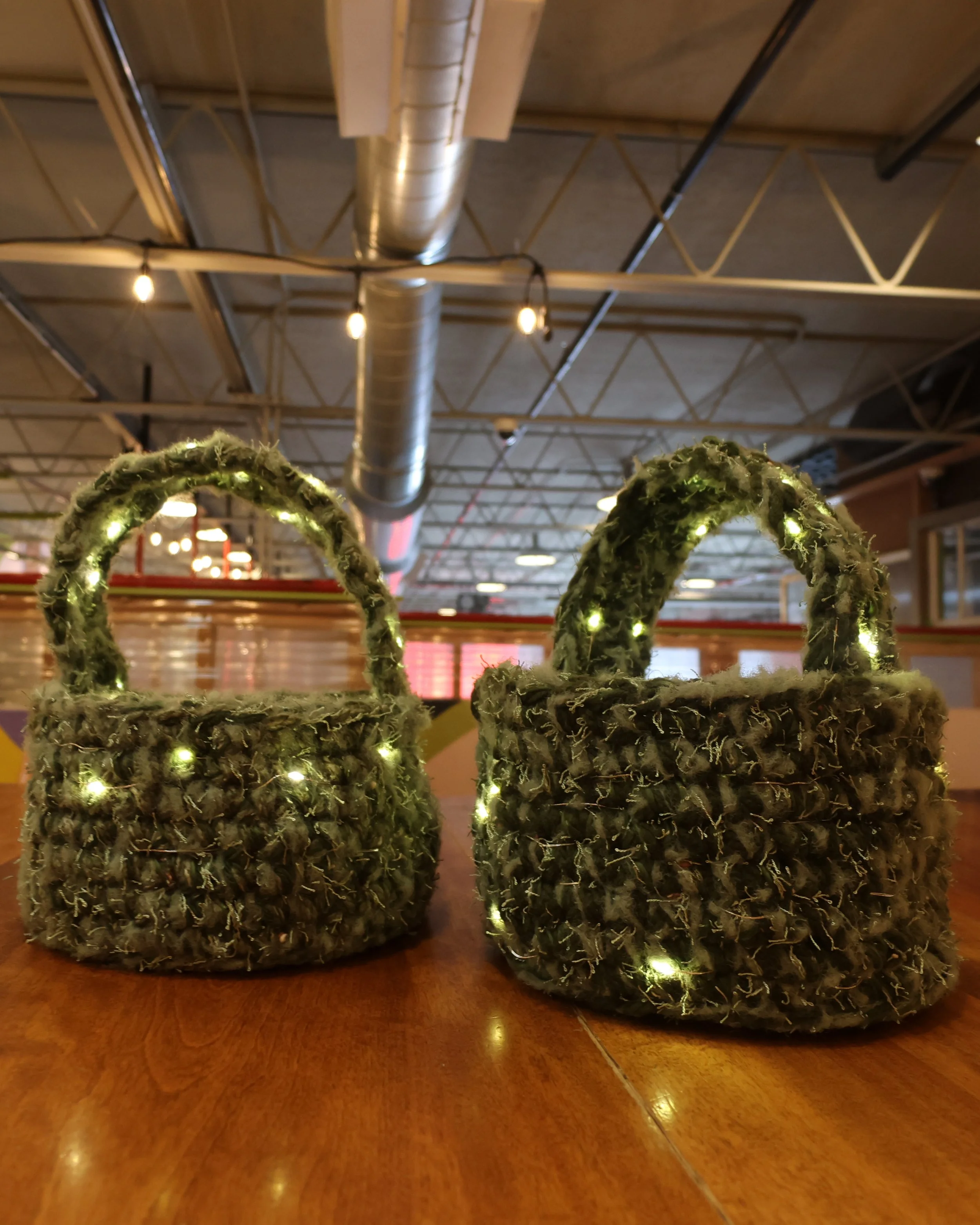 Close-up of two baskets made of moss-like yarn with tiny lights, sitting on a wooden surface, with an industrial ceiling with ductwork and exposed beams in the background.
