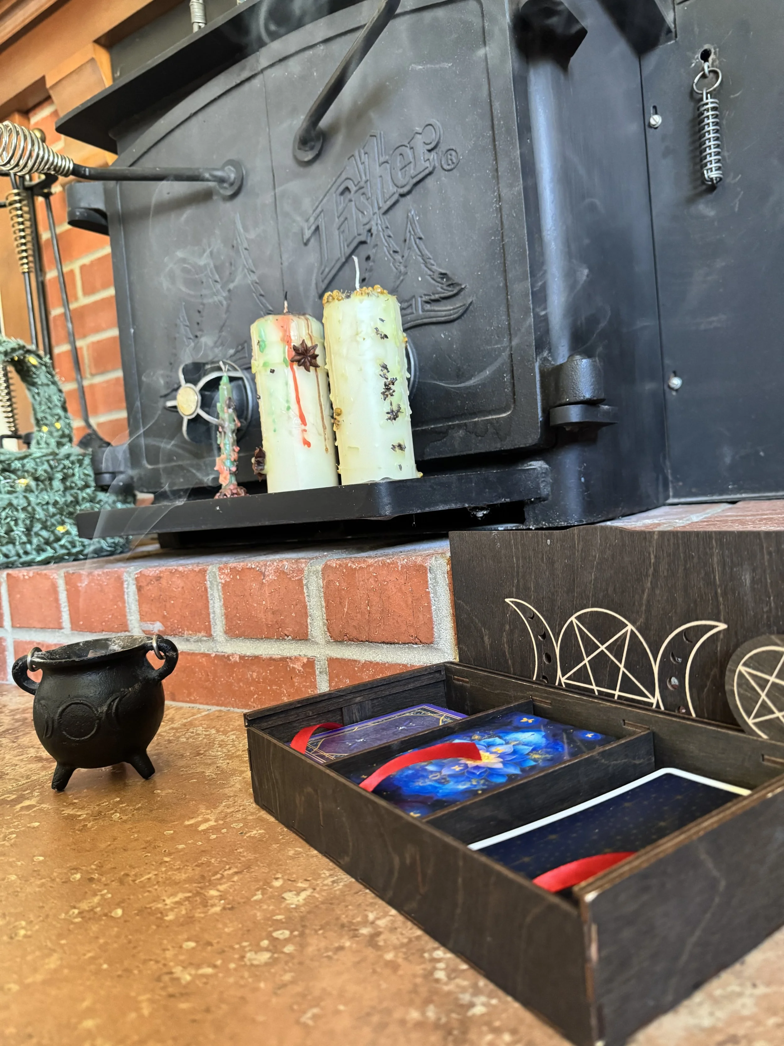 Three colorful candles with various decorations placed on a black metal fireplace. To the left, a black cauldron are on a brick mantle. A box with tarot cards and a red ribbon is partially visible in the foreground.