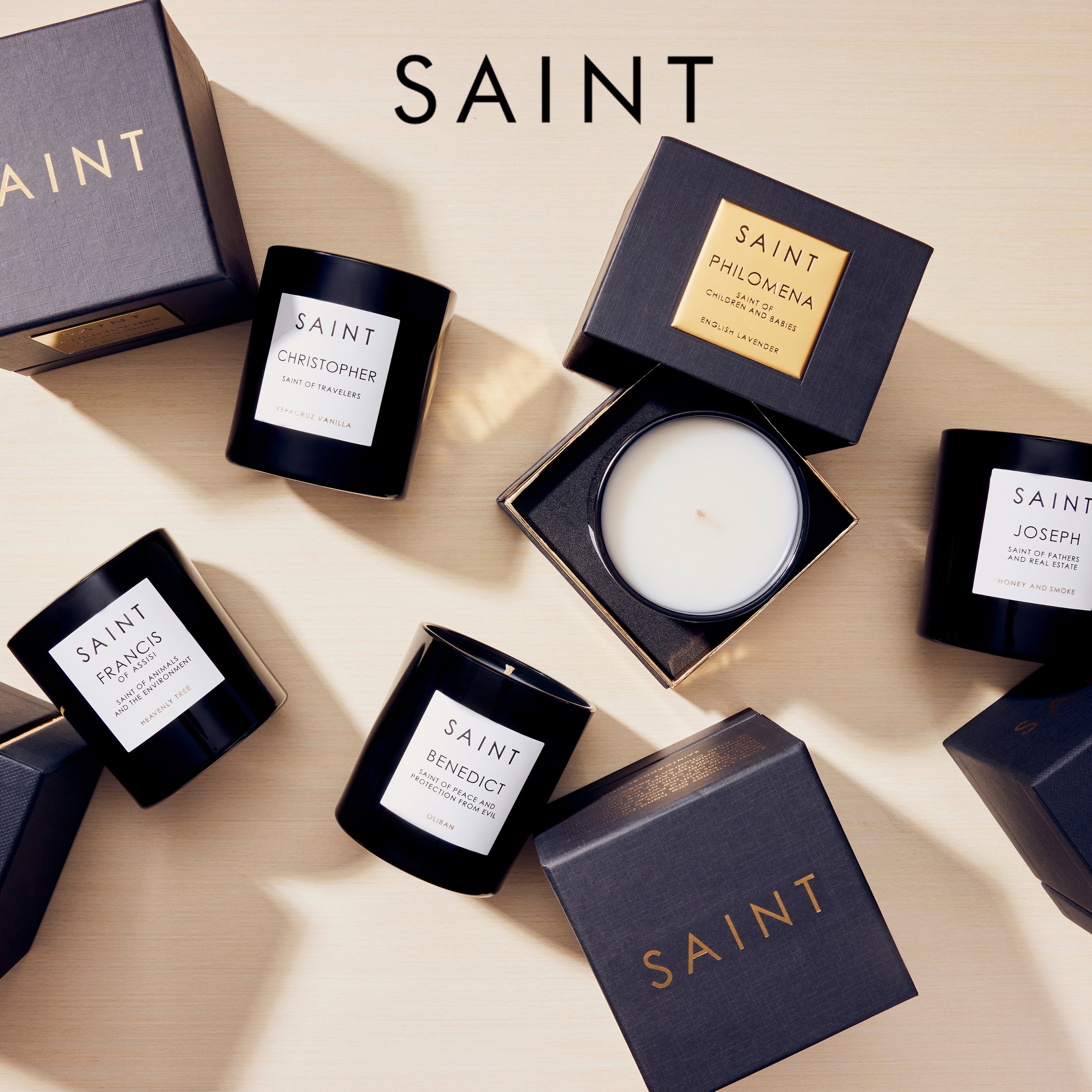Various scented candles and boxed gift sets labeled with saint names, arranged on a light wooden surface.