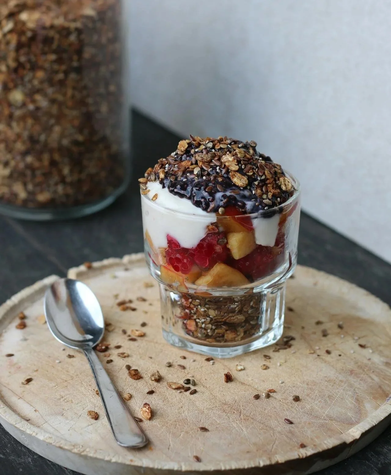 A layered yogurt parfait in a glass with granola, fresh fruit, and berry sauce, topped with granola and seeds, placed on a wooden tray with a spoon and scattered granola.