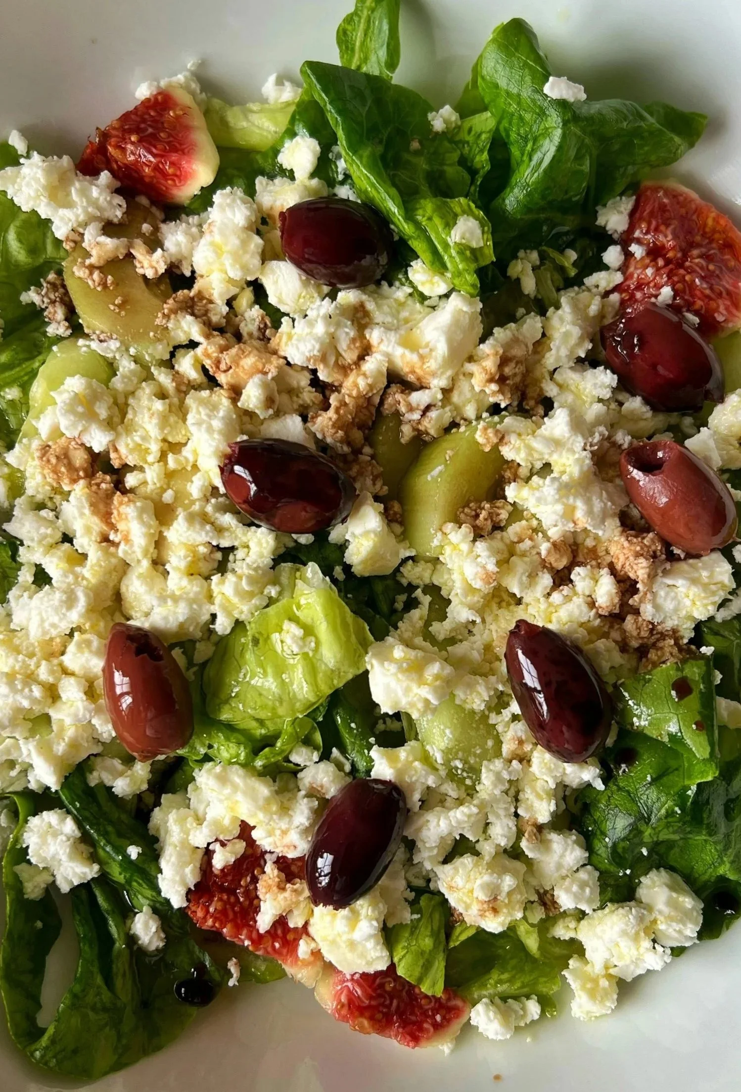 A fresh green salad with sliced figs, black grapes, crumbled feta cheese, and chopped walnuts on a white plate.