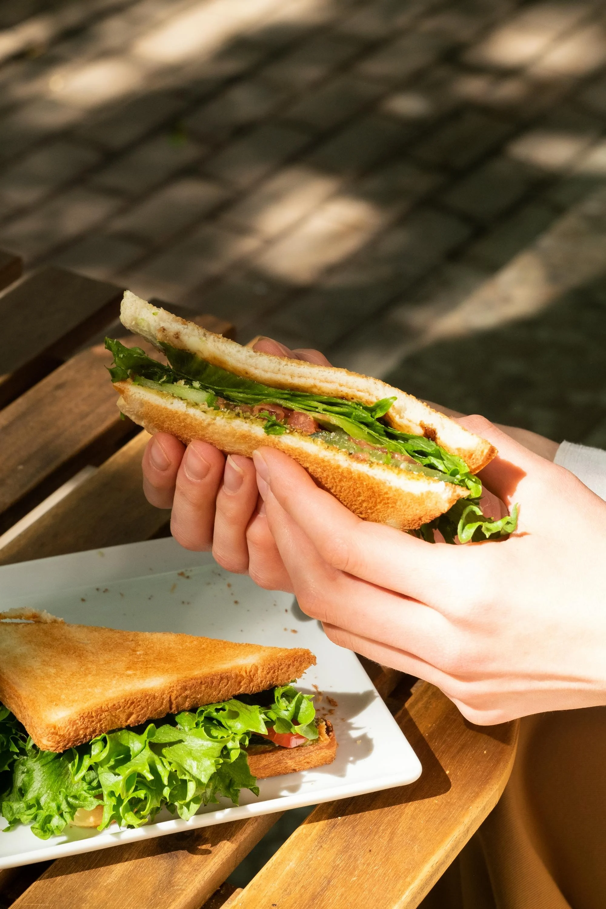 Person holding a sandwich with lettuce, ham, and tomato, with another sandwich on a white plate on a wooden table.