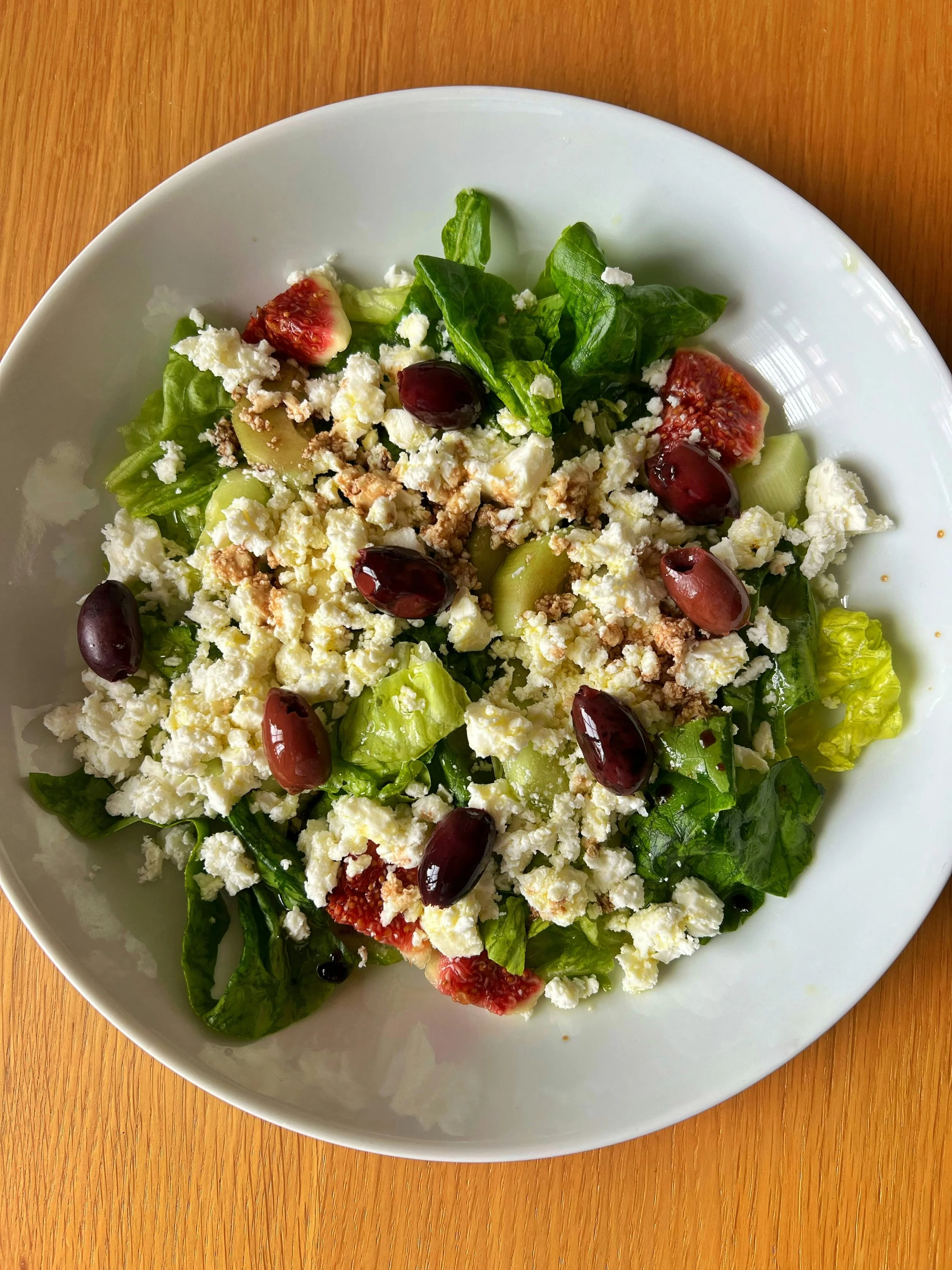 A plate of fresh salad with lettuce, spinach, strawberries, black olives, feta cheese, chopped walnuts, and sliced cucumbers on a wooden surface.