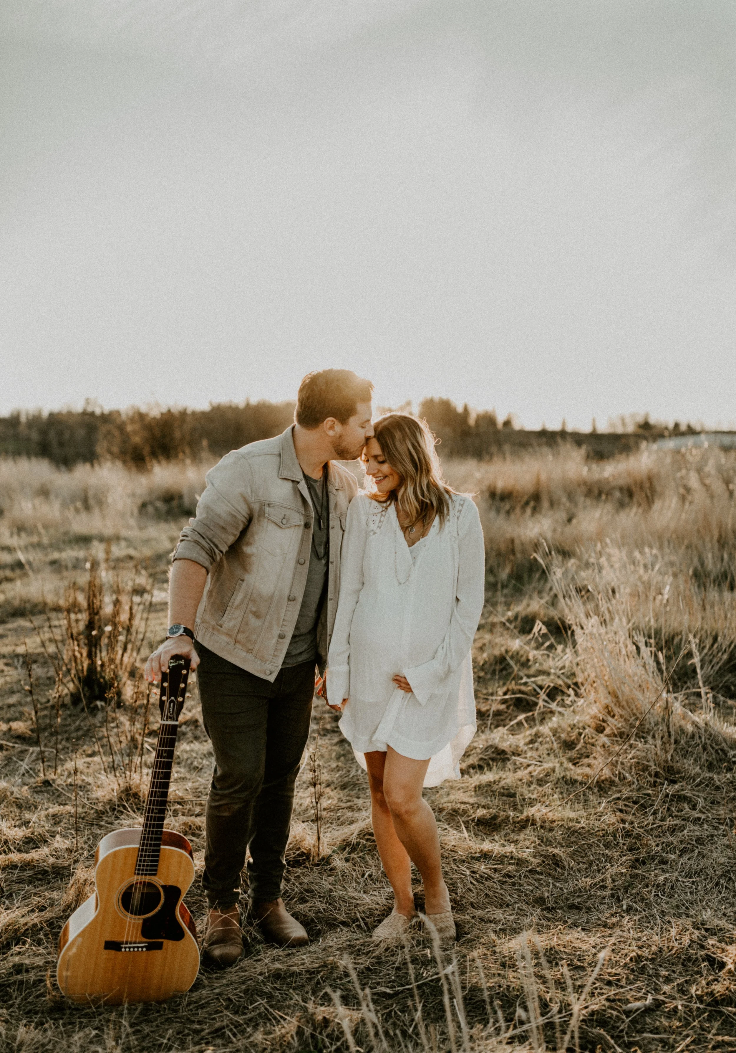 A young man and woman share a tender moment in a field during sunset, with the man gently kissing the woman's forehead and a guitar resting on the ground nearby.