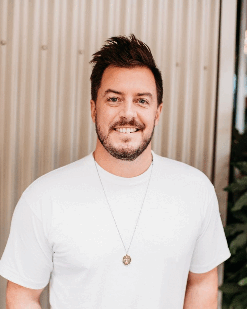 A smiling man in a white t-shirt and necklace standing in front of a corrugated metal wall.