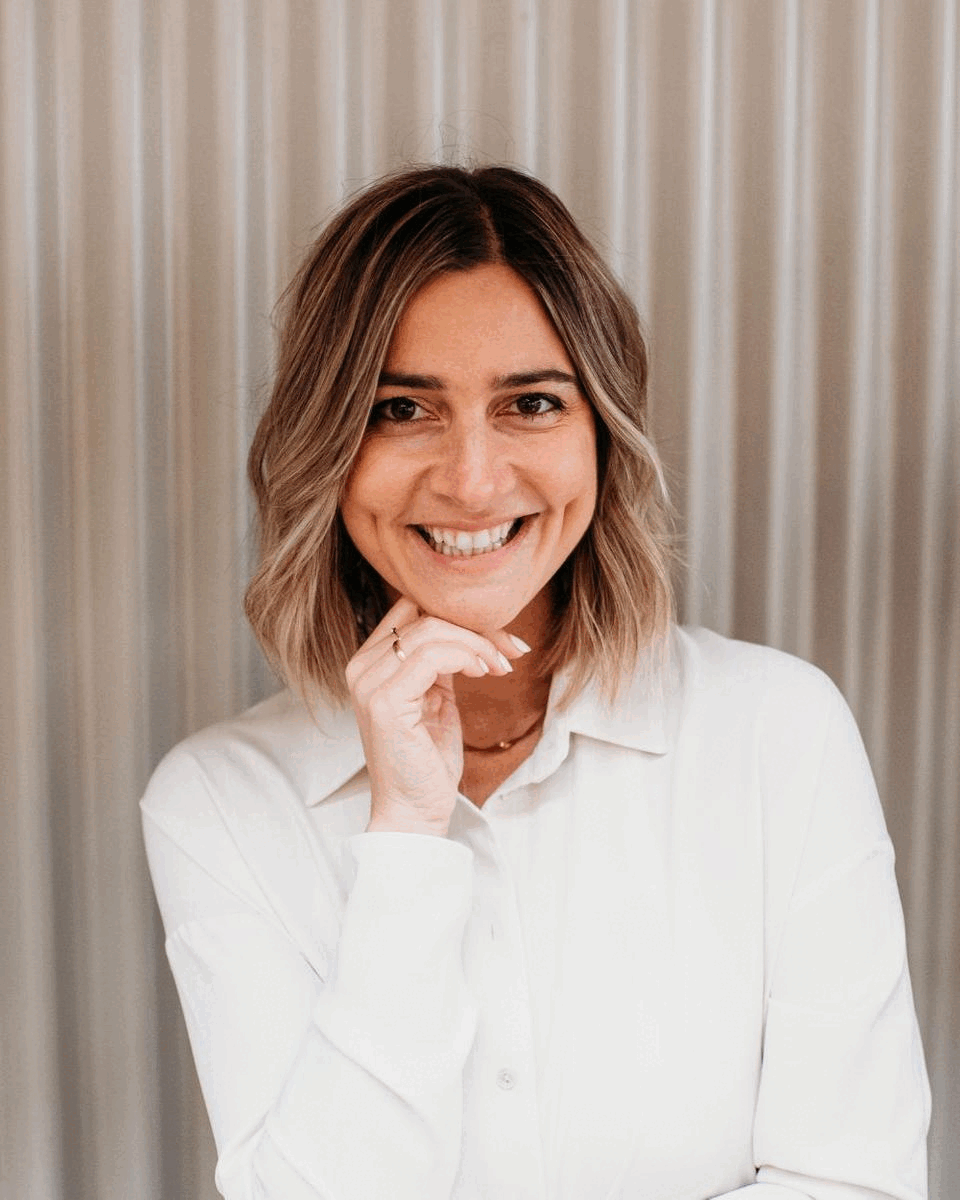 A woman with shoulder-length brown hair smiling, dressed in a white shirt, posing in front of light-colored curtains.
