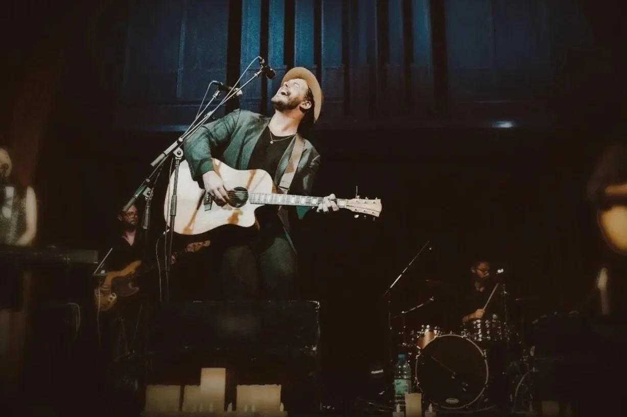 Musician playing guitar and singing on stage with band members in the background.