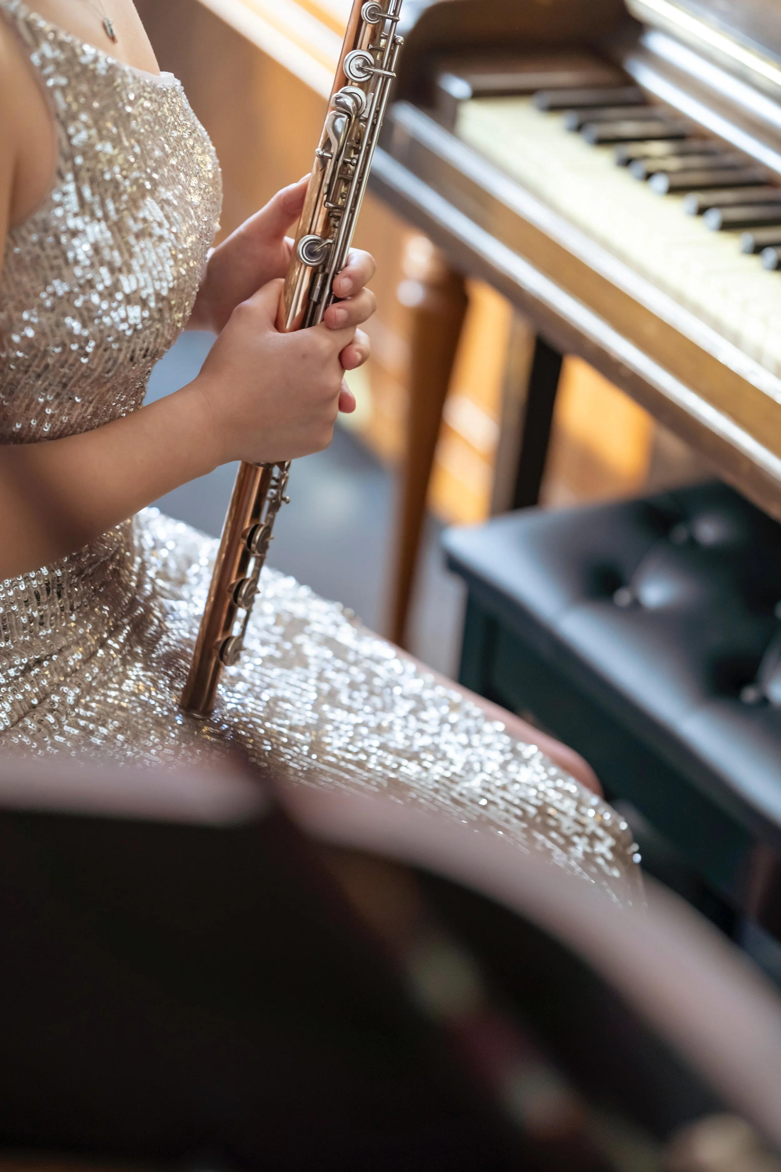 Person in a sparkly, gold dress playing a silver flute near a piano.