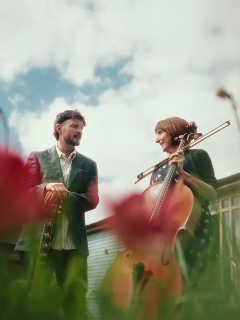 A man and a woman are outdoors, talking, with the woman playing a cello and the man holding a guitar. The sky is partly cloudy.