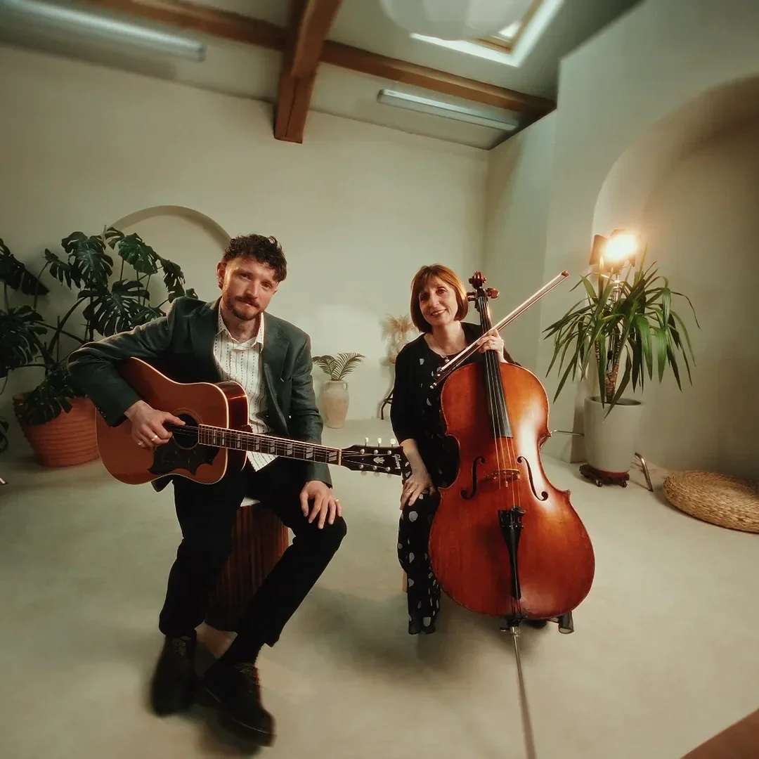 A man playing guitar and a woman playing cello in a room with plants and a skylight