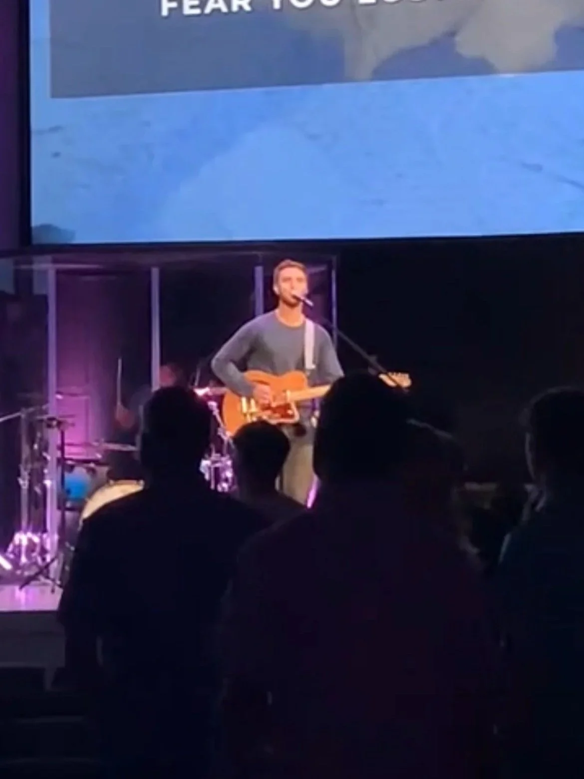 A man is performing on a stage, playing an acoustic guitar and singing into a microphone, with an audience in front of him. He can play shows at corporate events or private parties as well.