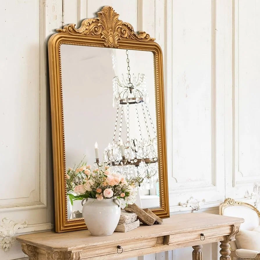 Elegant mirror with an ornate gold frame, placed on a wooden console table decorated with a white vase of pink and white roses, a small stack of books, and a decorative wooden piece, all set against a light-colored paneled wall.