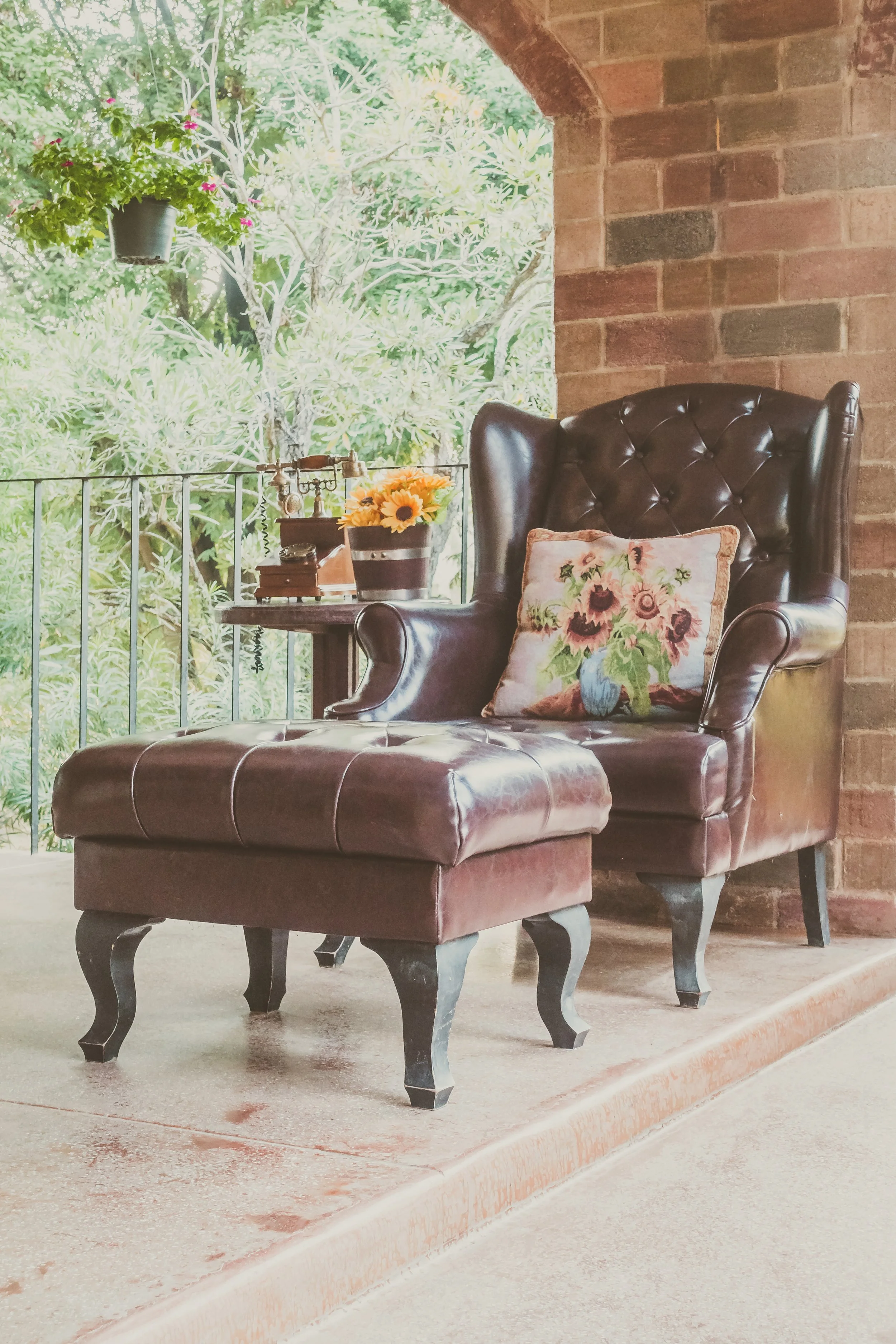 A leather armchair with a floral pillow, a matching ottoman, a small round wooden table with a bouquet of sunflowers, a vintage telephone, and a bronze box, set on a porch with a brick wall and lush greenery in the background.