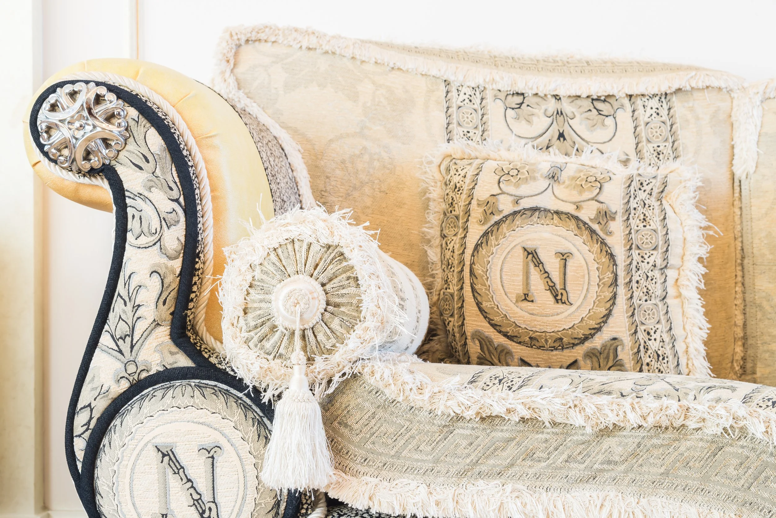 Close-up of ornate beige and cream furniture with embroidered patterns, including a pillow with frayed edges, a decorative tassel, and a floral silver embellishment.
