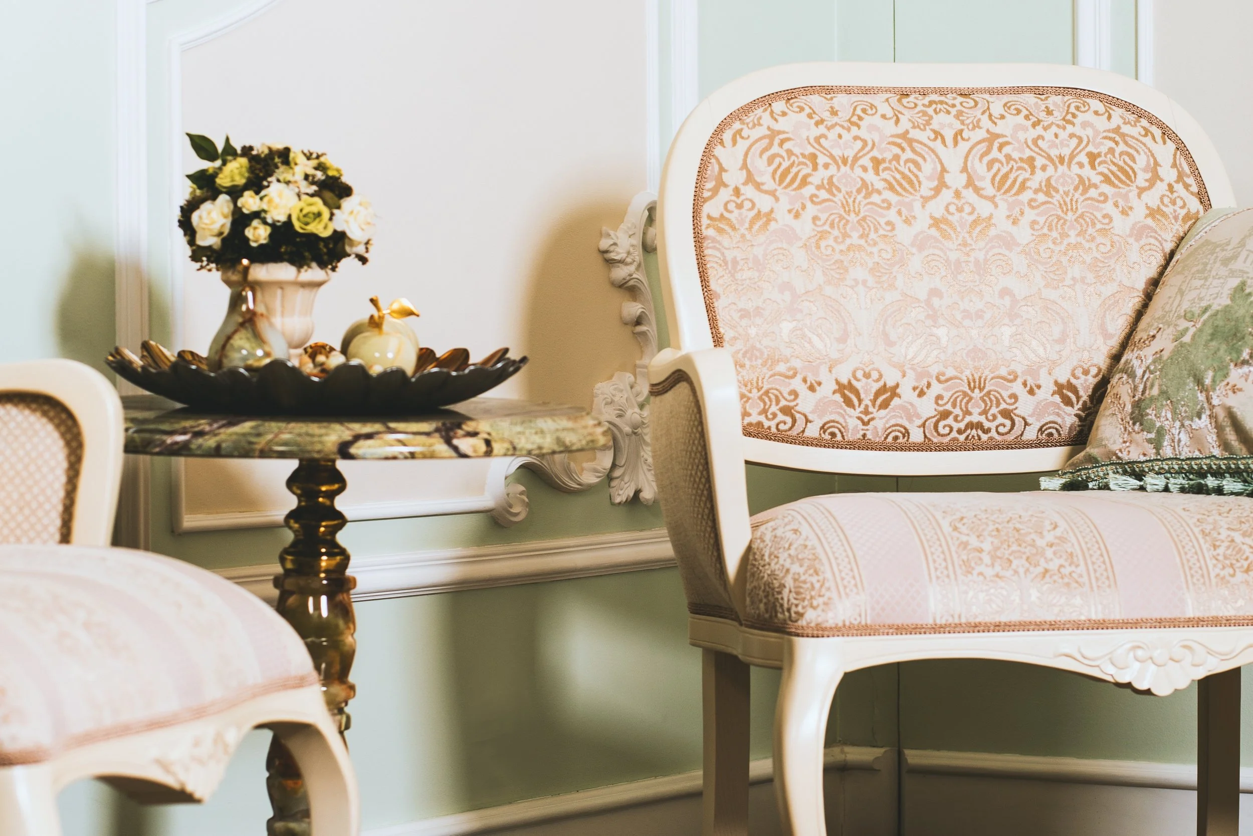 A vintage-style white wooden chair with ornate carvings and patterned pink upholstery, positioned in a light green room with white wainscoting, next to a small round table with a marble top and decorative floral arrangements.
