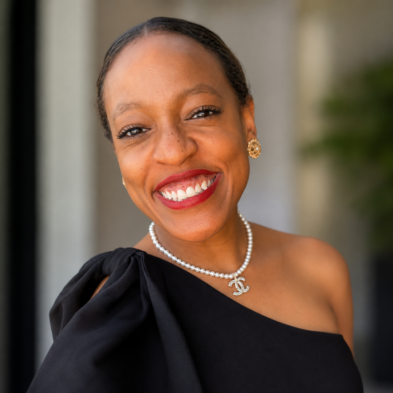 A woman with dark hair in a bun, wearing a black dress with a single shoulder strap, a pearl necklace with a Chanel logo pendant, matching pearl earrings, and red lipstick, smiling at the camera.