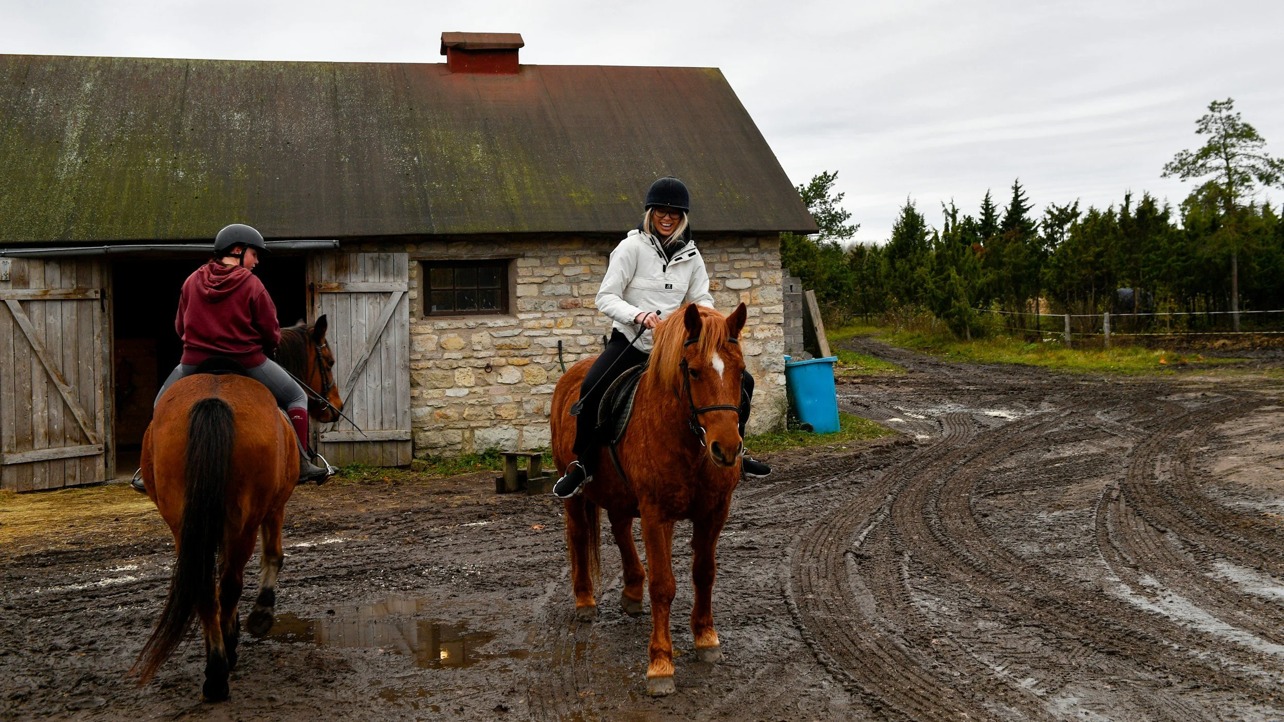 Two people riding horses near a rustic stone and wood barn on a muddy farm path under cloudy skies.
