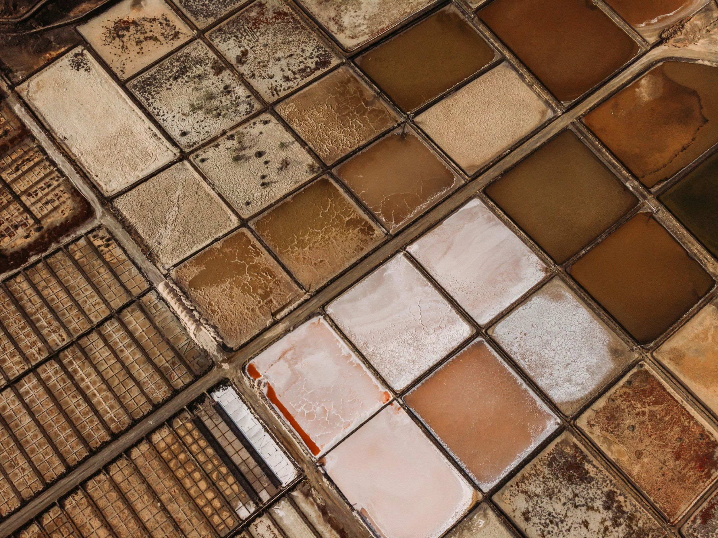Aerial view of salt evaporation ponds with various shades of brown, beige, and white, separated by narrow embankments.