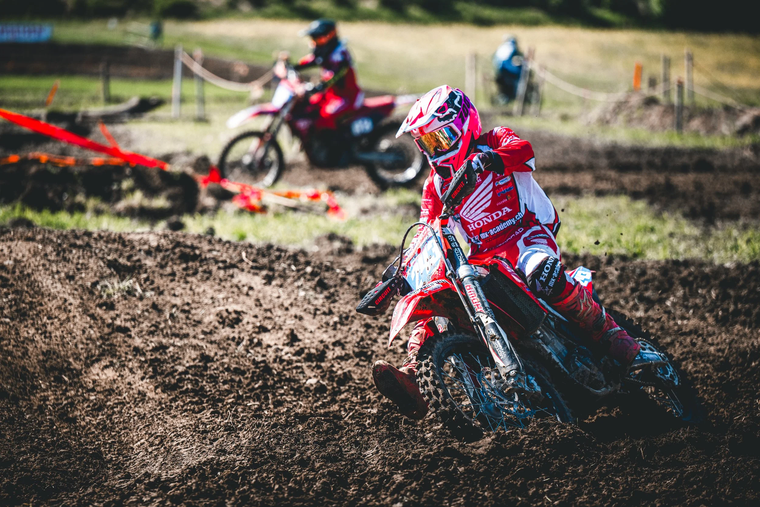 Ein Honda-Motocross-Fahrer in roter Gear und auffälligem Helm zieht durch eine schlammige Kurve beim SAM-Rennen in Hünikon (Thurgau), dynamische Schweizer Sportfotografie.