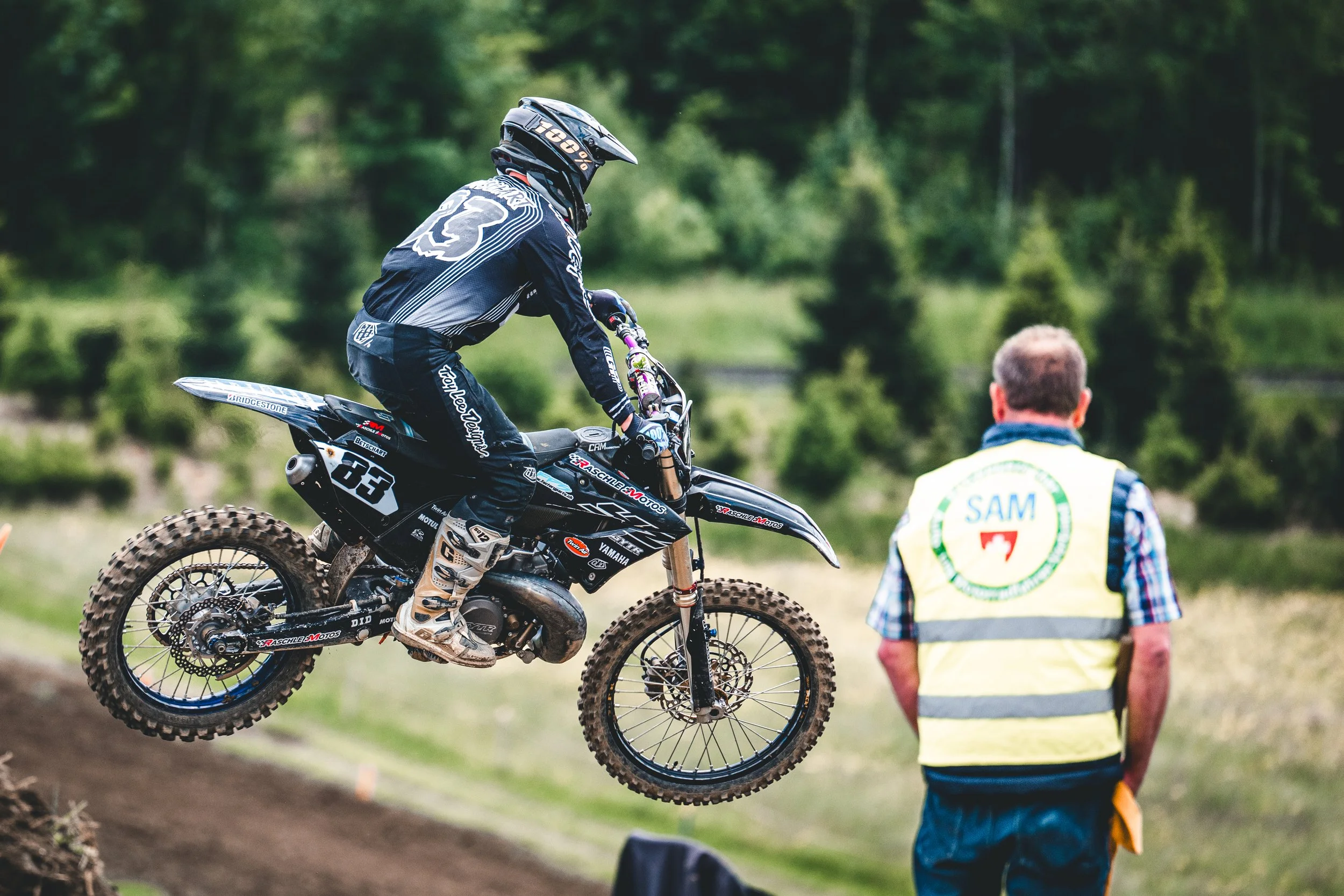 Ein Motocross-Fahrer mit der Startnummer 83 im weiten Sprung, beobachtet von einem SAM-Streckenposten in Hünikon (Thurgau), authentische Sportreportage aus der Schweiz.