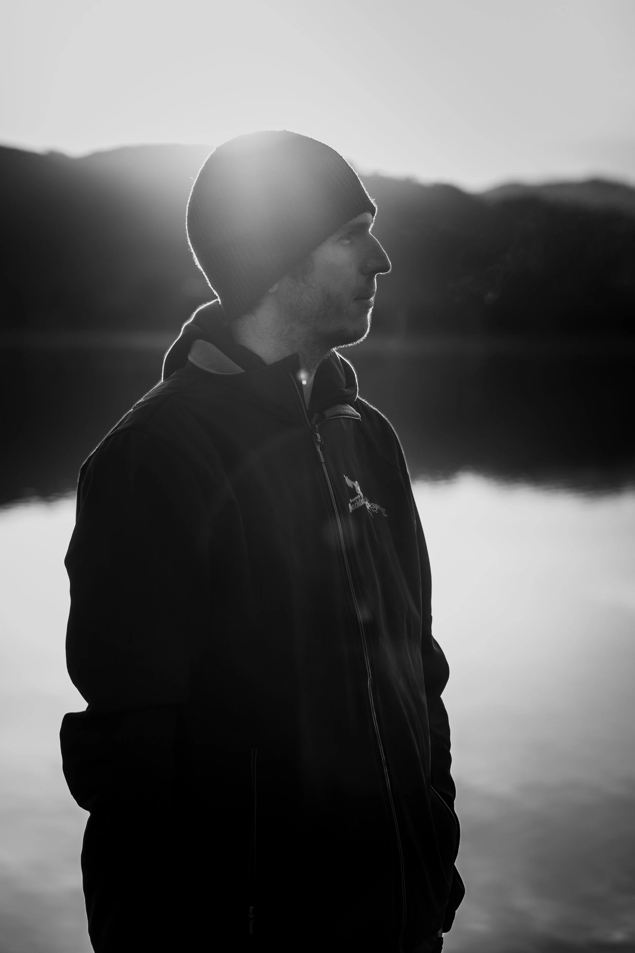 Mann steht am Wasser bei Sonnenuntergang, trägt eine schwarze Jacke und eine Mütze, Blick nach rechts, schwarz-weiß
