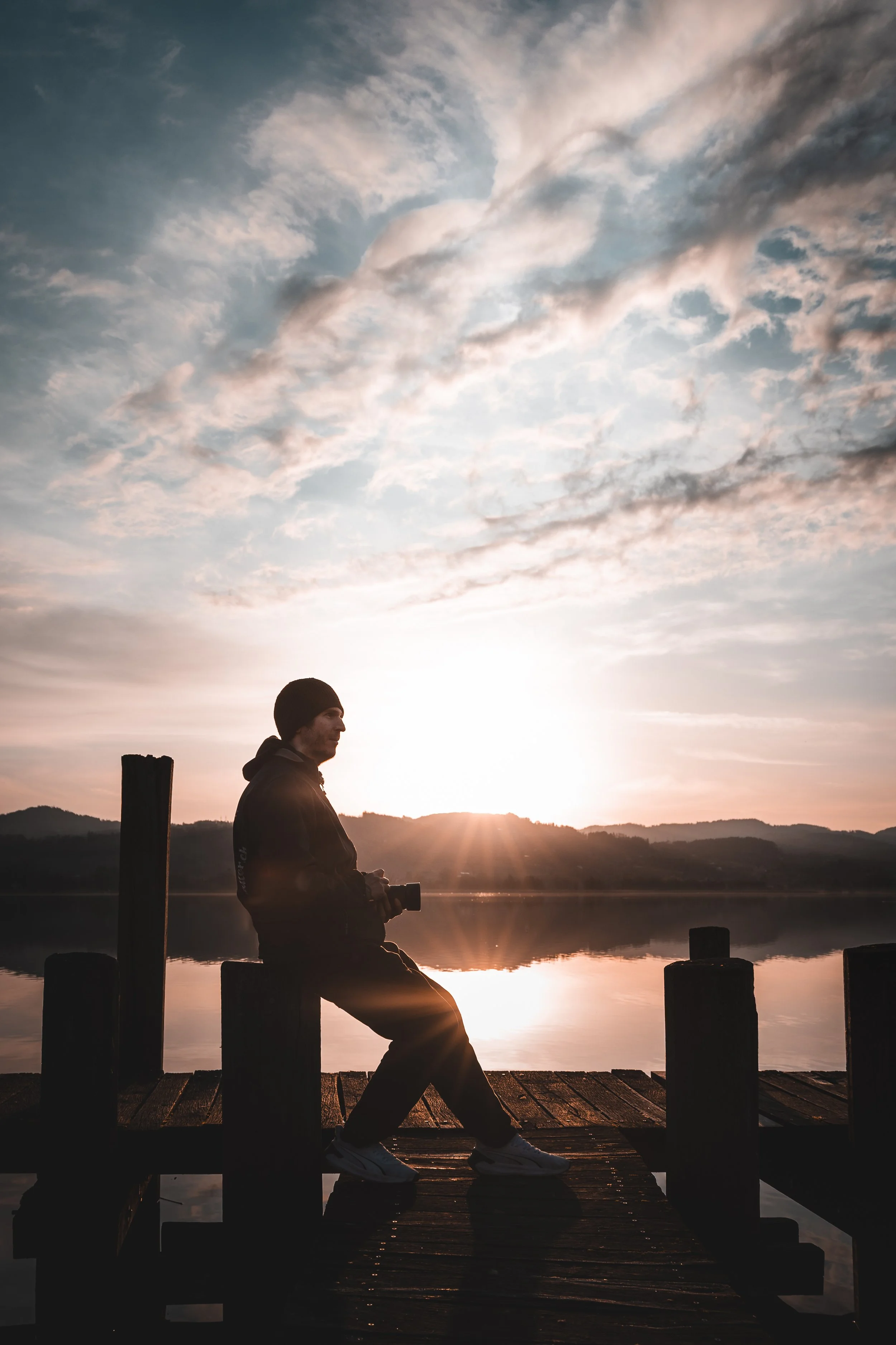 Eine Person sitzt auf einem Steg am See bei Sonnenuntergang, hält eine Kamera in den Händen, trägt eine schwarze Mütze und Hoodie, im Hintergrund sind Bergketten und ein bewölkter Himmel sichtbar.