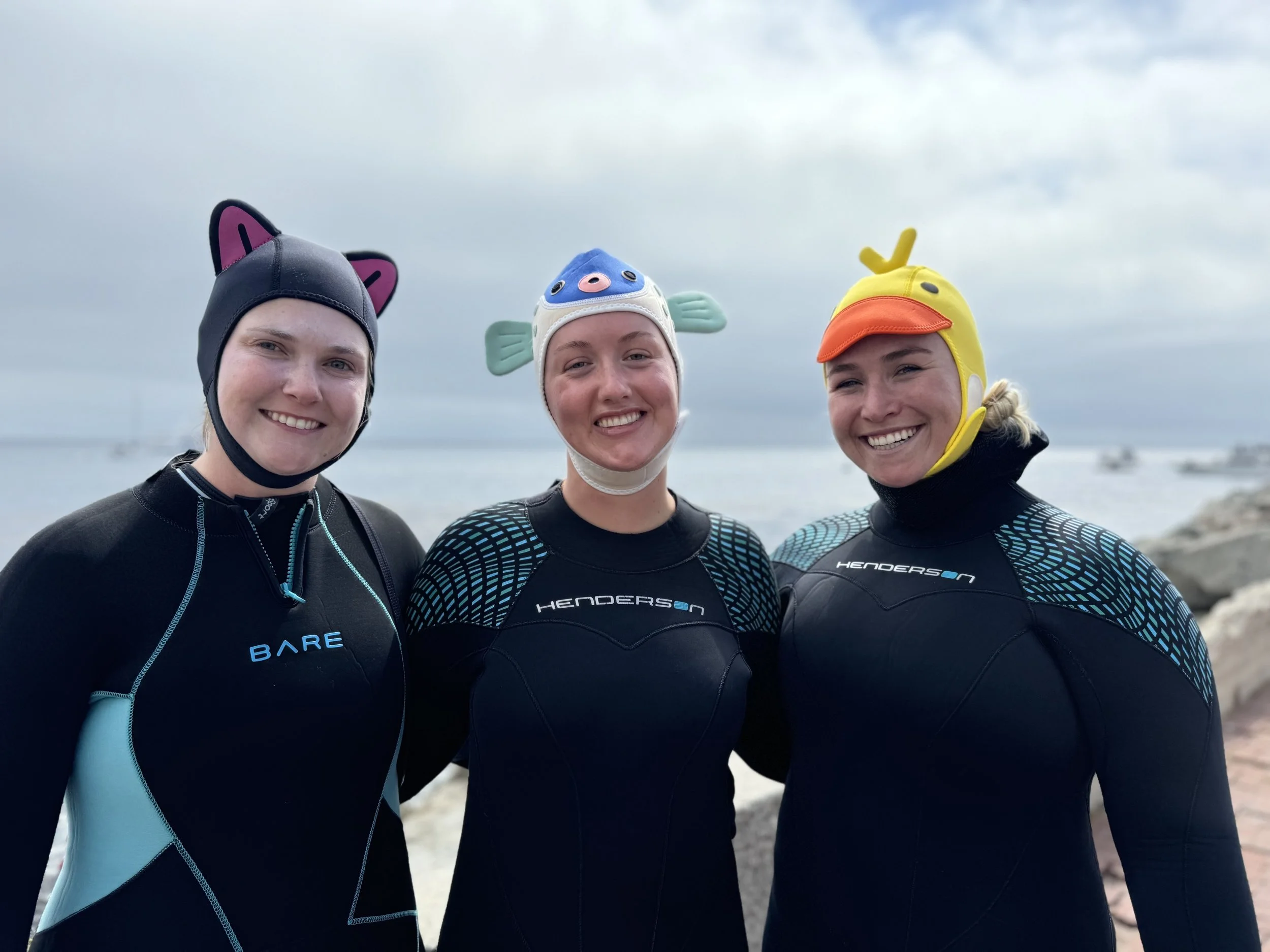 Three smiling women at the beach wearing wetsuits and unique animal-themed swim caps, with a cloudy sky and ocean in the background.