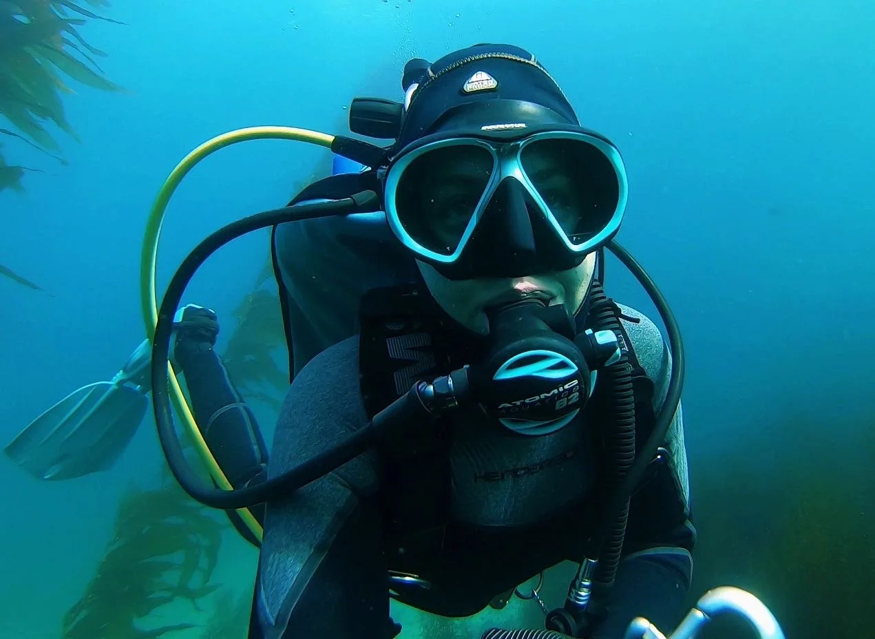 A person scuba diving underwater wearing a mask, regulator, and wetsuit with a yellow air hose, surrounded by blue water.