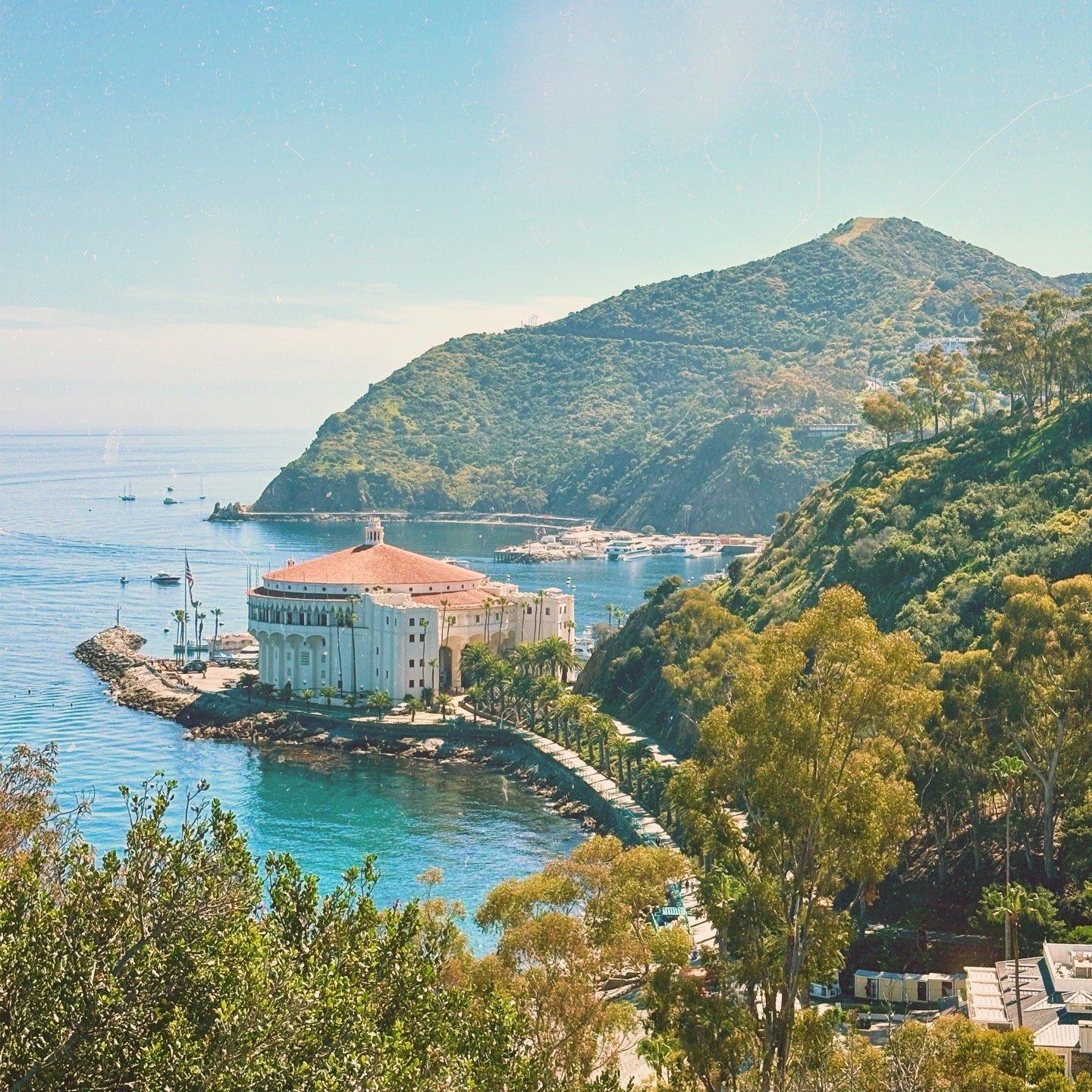 Coastal scene with a large white building with a red roof on a small peninsula, green hills in the background, and boats on the water.