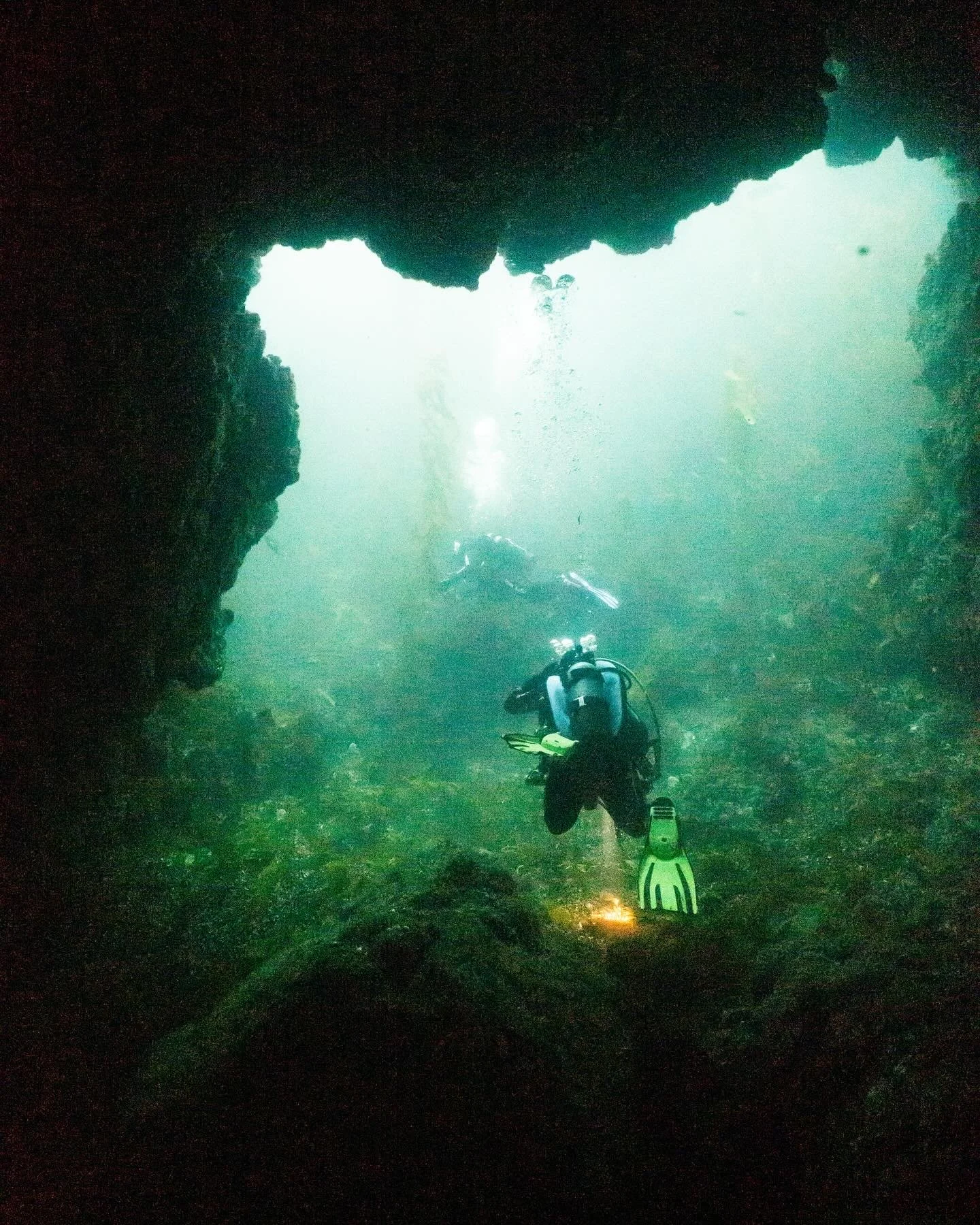 Underwater cave with a scuba diver swimming near the bottom and sunlight shining through an opening in the rocks.