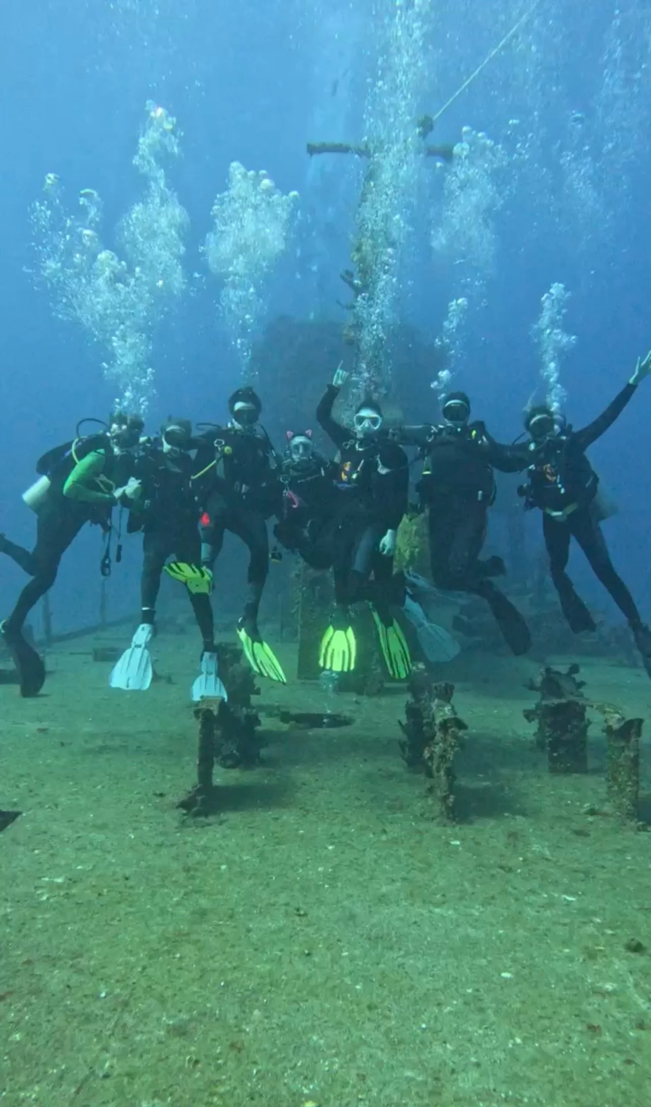 A group of scuba divers underwater in a clear blue ocean, wearing wetsuits, masks, and flippers, positioned on a shipwreck or submerged structure.
