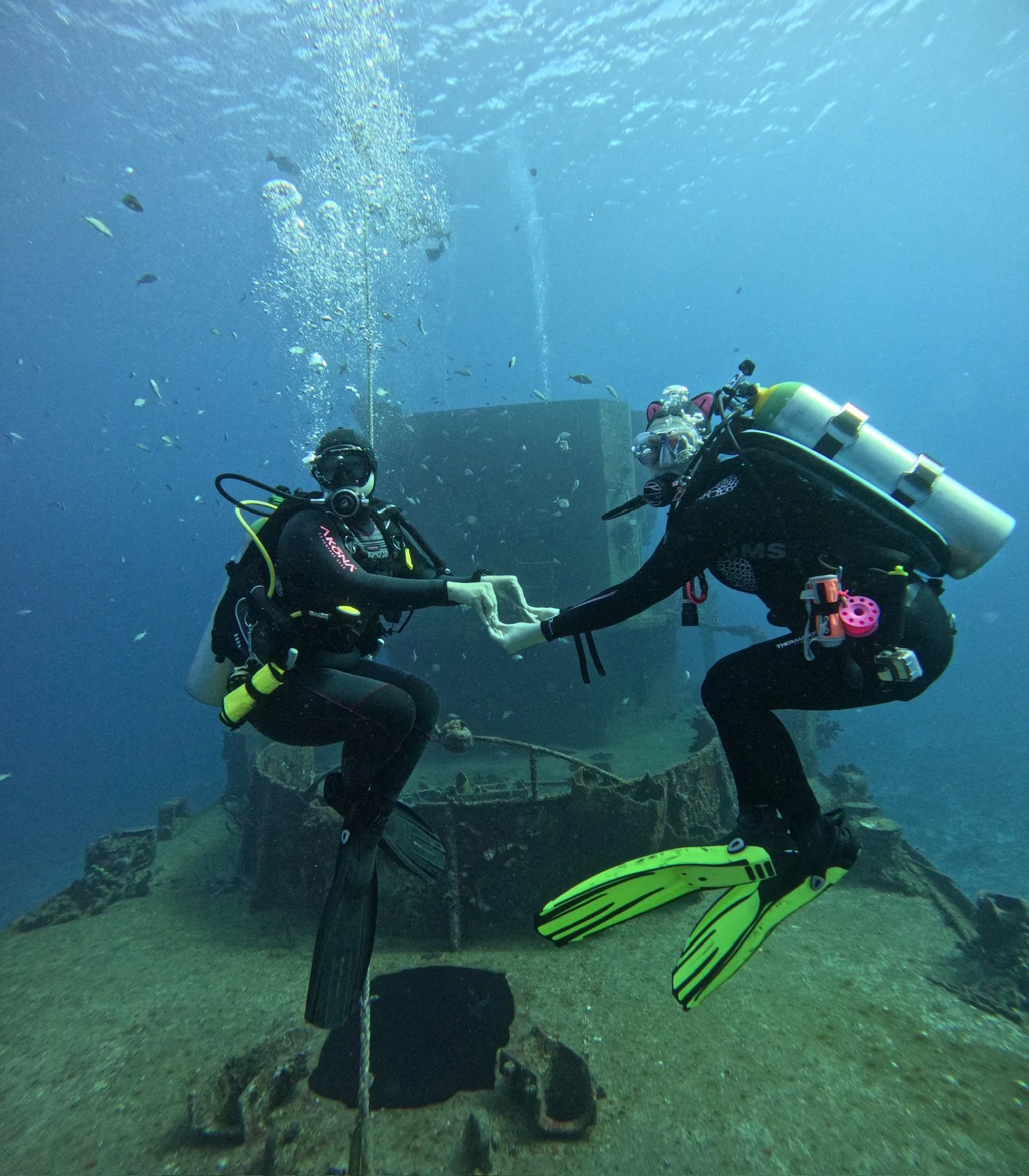 Underwater scene with scuba divers holding hands, surrounded by small fish and bubbles, with a submerged structure and sea floor visible.