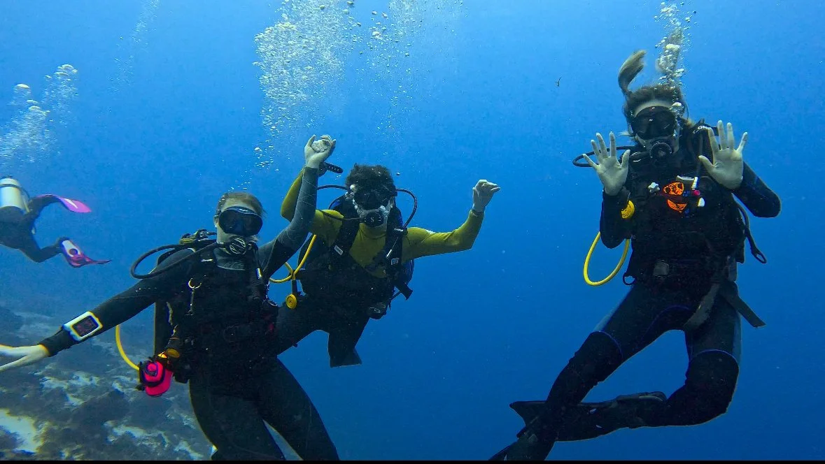Three scuba divers underwater, posing with hand signals and surrounded by blue ocean water.