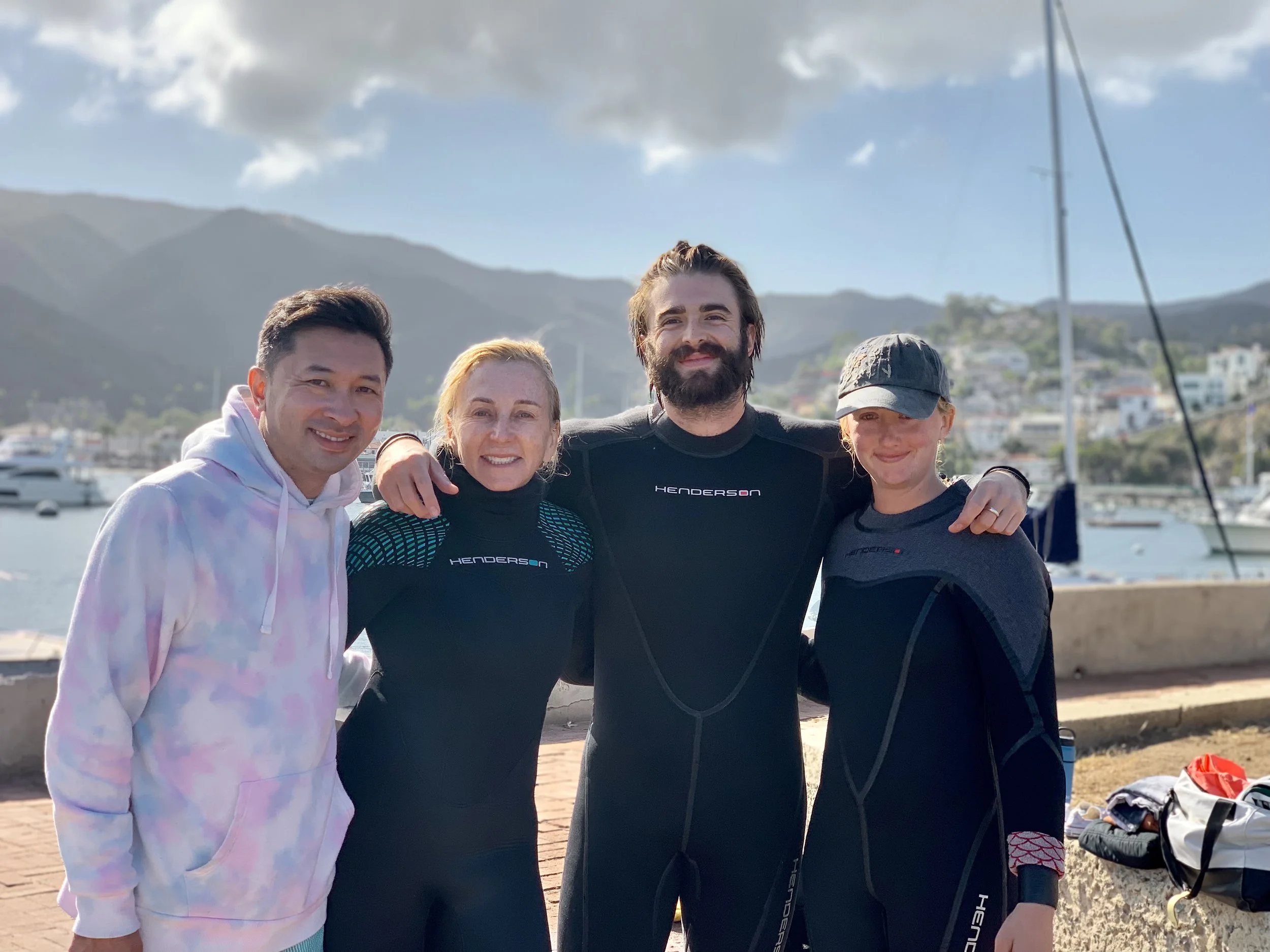 A group of four smiling people, two men and two women, standing together outdoors near the water with a marina and mountains in the background. They are dressed casually, some in athletic wear, with a sailboat mast visible on the right.