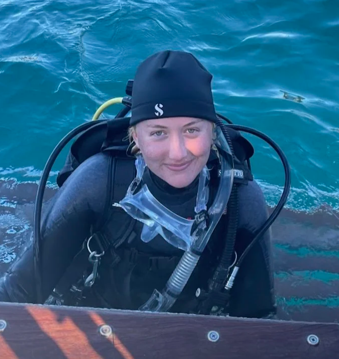 Person in scuba gear with a black beanie, smiling, in the water, near a boat.