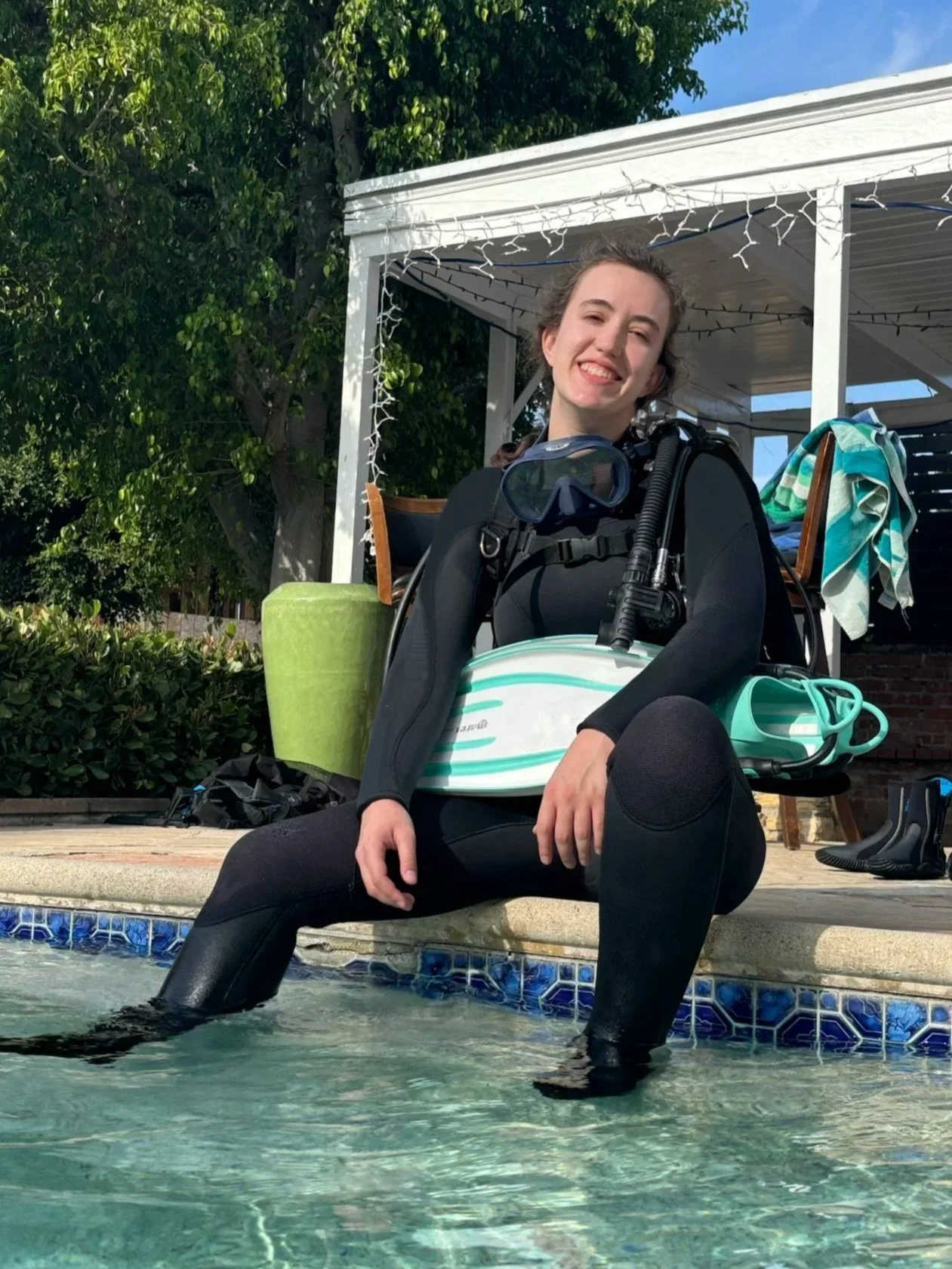 A young woman sitting on the edge of a swimming pool, smiling, wearing a wetsuit, with snorkeling gear on her chest, and her feet submerged in the water. Behind her are poolside items, a covered patio with string lights, and lush greenery.