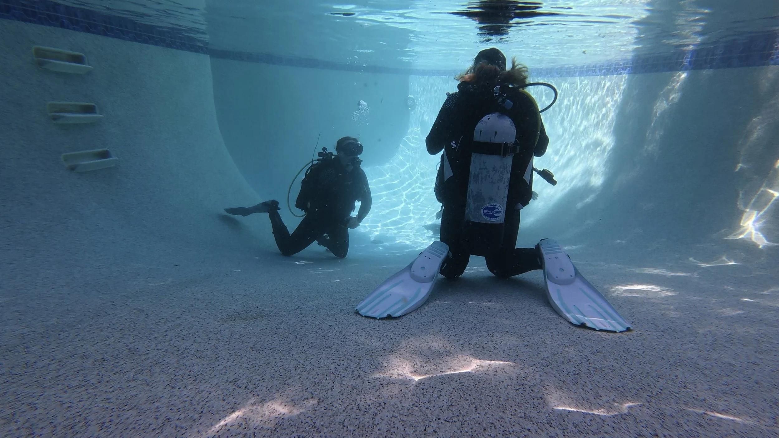Two scuba divers underwater in a swimming pool, one kneeling with fins and the other standing with fins, surrounded by blue water and pool walls.