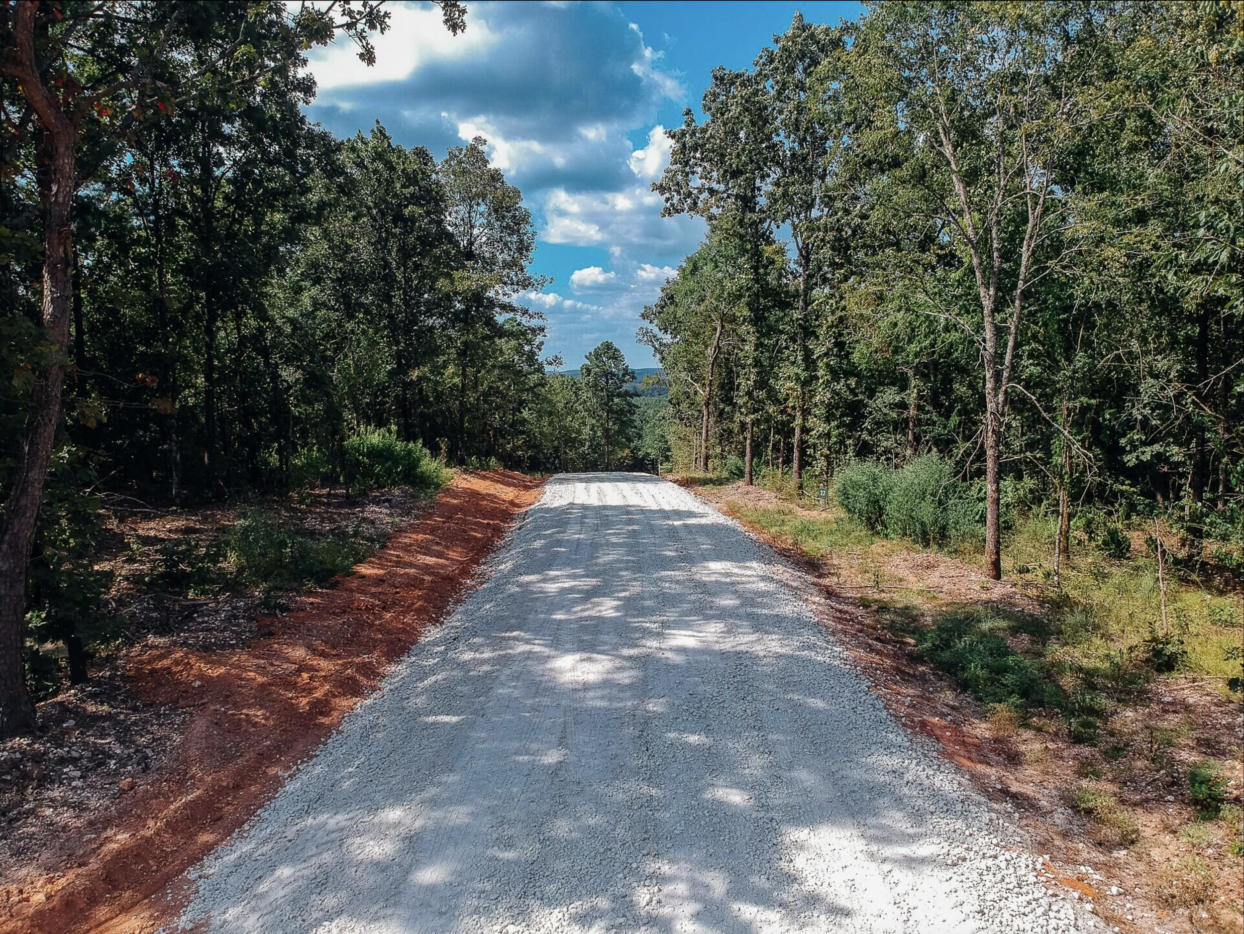 A gravel road runs through a forested area with trees on both sides under a partly cloudy sky.